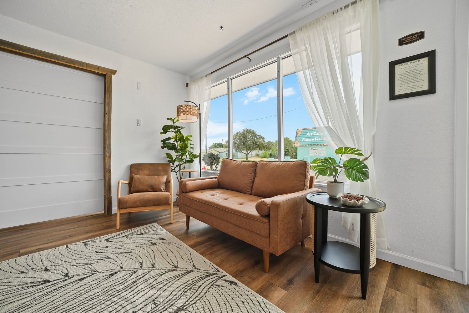 Bright living room with brown leather sofa, wooden armchair, large window with sheer curtains, and hardwood floors.