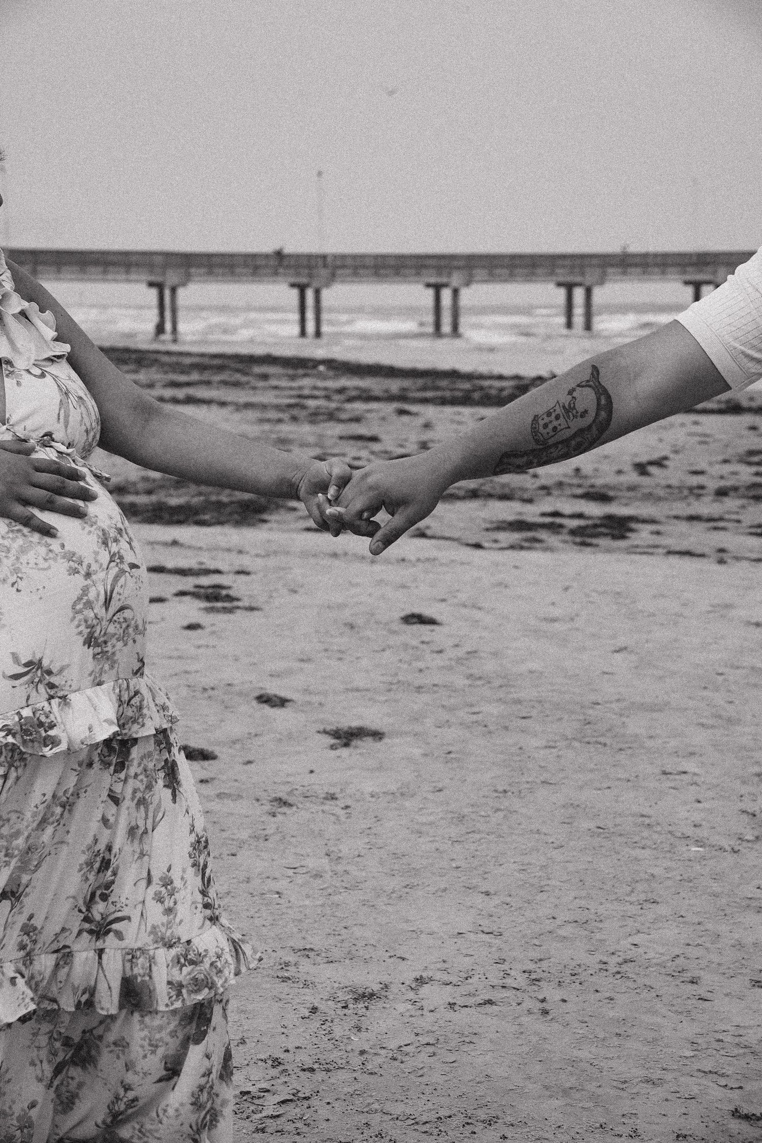 Black and white photo of a couple holding hands on a beach, man's tattooed arm visible, pier in background.