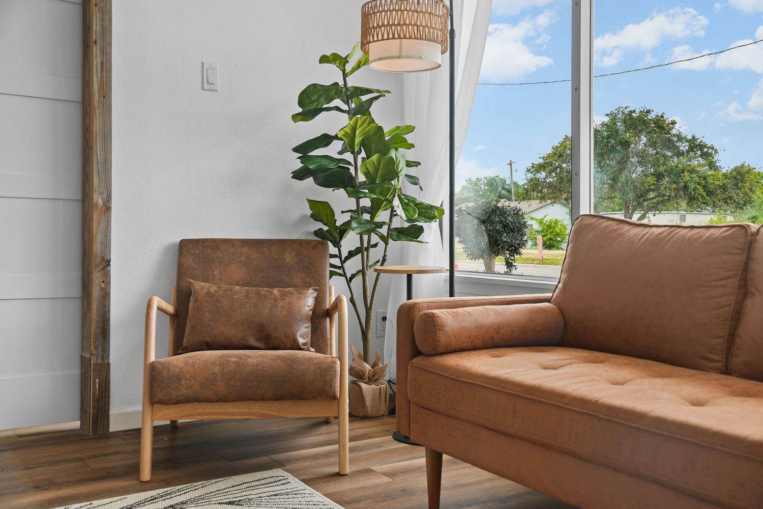 Sunlit sitting area with brown leather sofa, matching armchair, fiddle leaf fig plant, and large corner window.