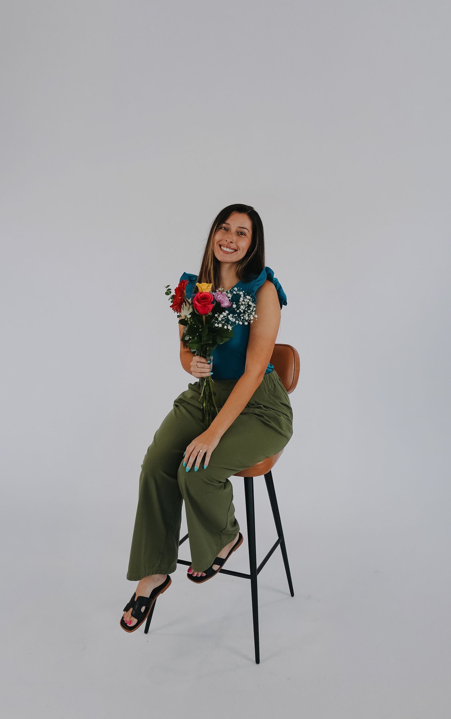 Smiling woman sitting on a stool holding colorful roses, wearing green pants and a teal top.