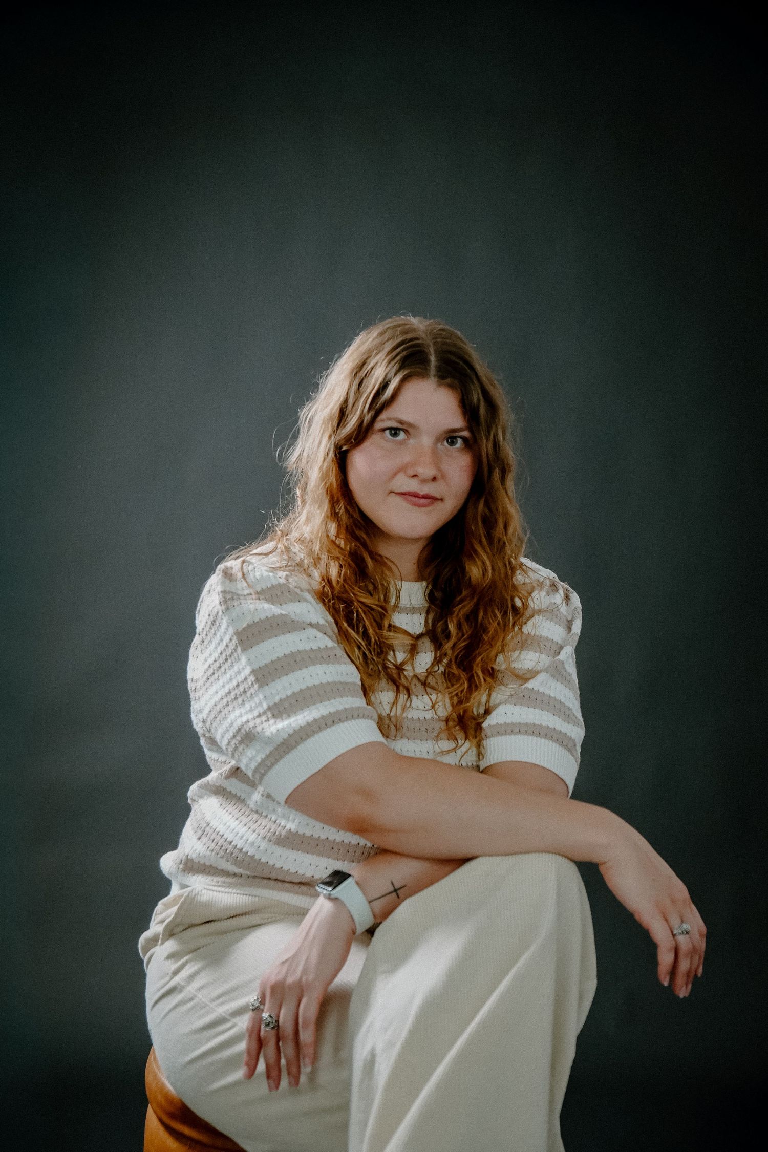 Young woman with curly auburn hair sits relaxed in studio, wearing striped sweater and white pants.