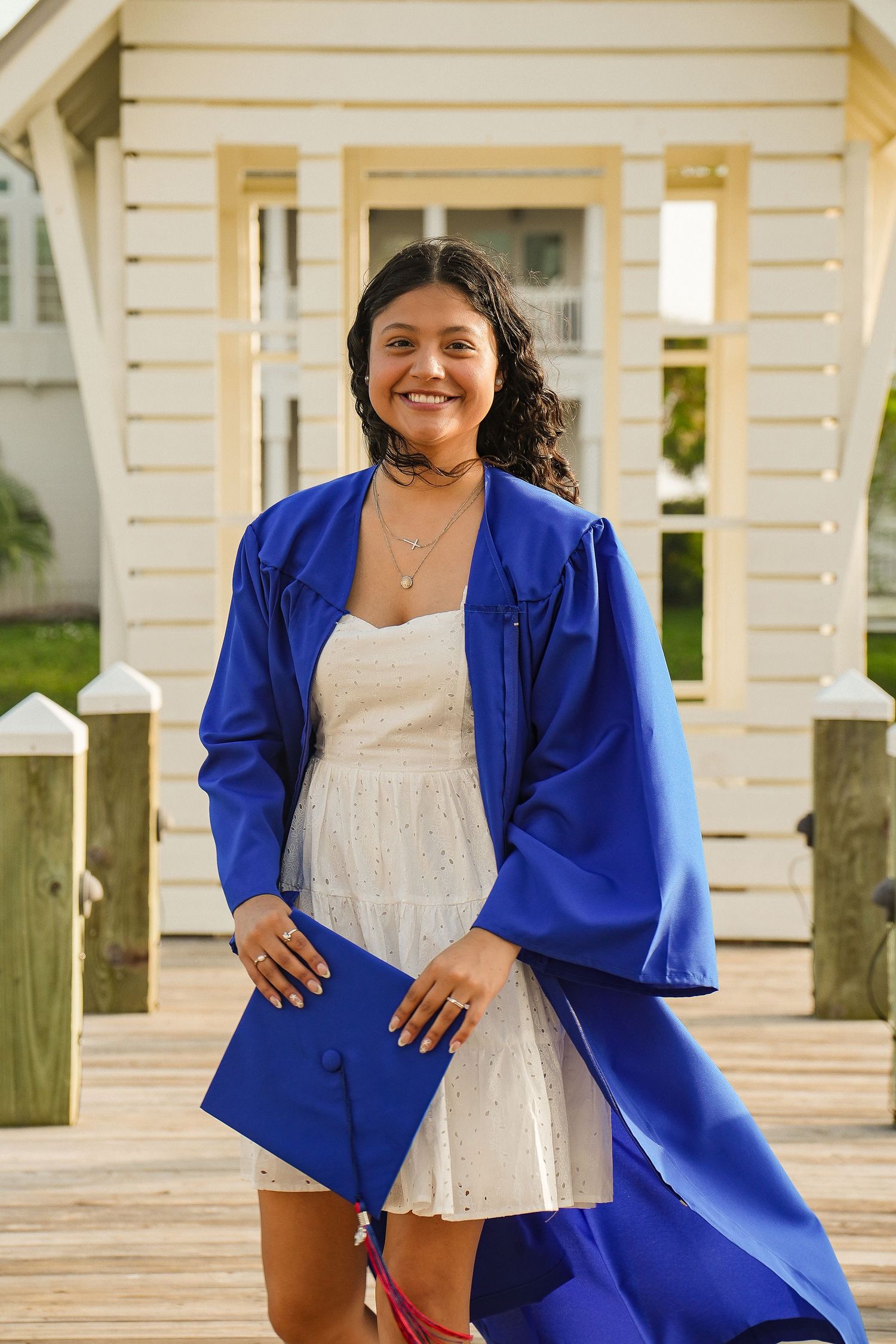 Smiling young woman in blue graduation gown and white dress holds cap on a sunny wooden deck.