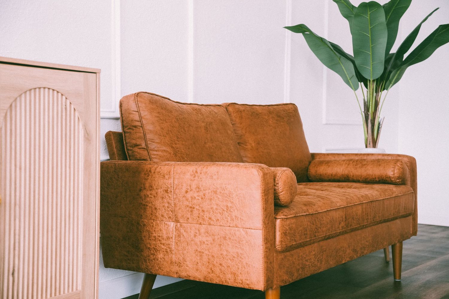 Distressed cognac leather loveseat with wooden legs beside a tropical plant against a white paneled wall.