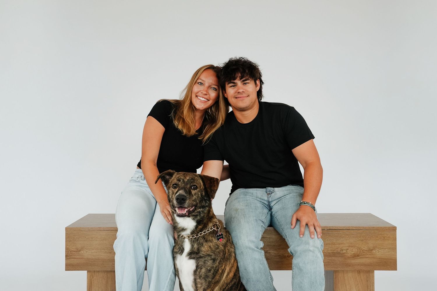 Two people in black shirts and jeans sit on a wooden bench with a brindle dog, smiling against a white background.