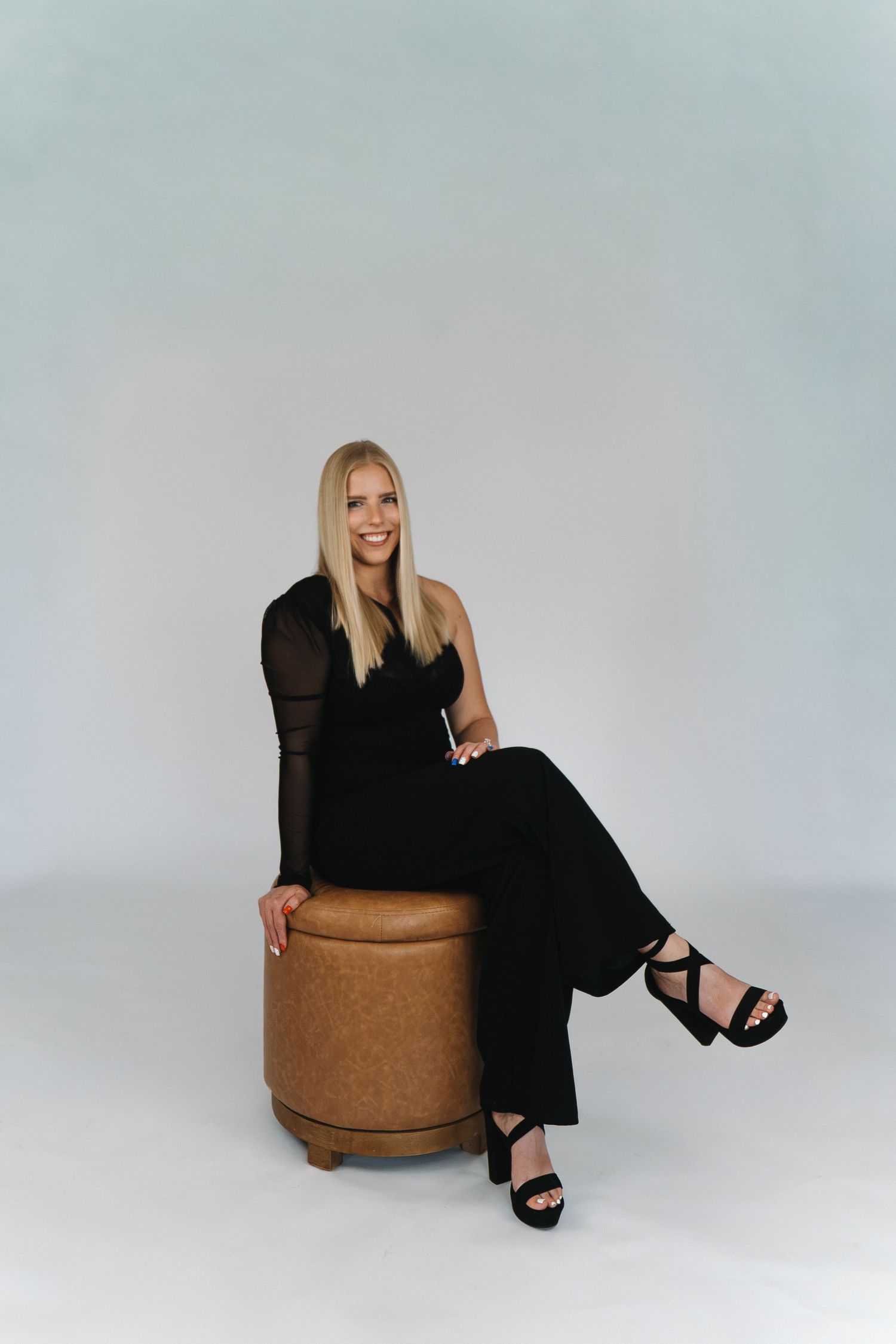 Blonde woman in black jumpsuit and heels sitting on a brown ottoman, smiling against a light gray background.
