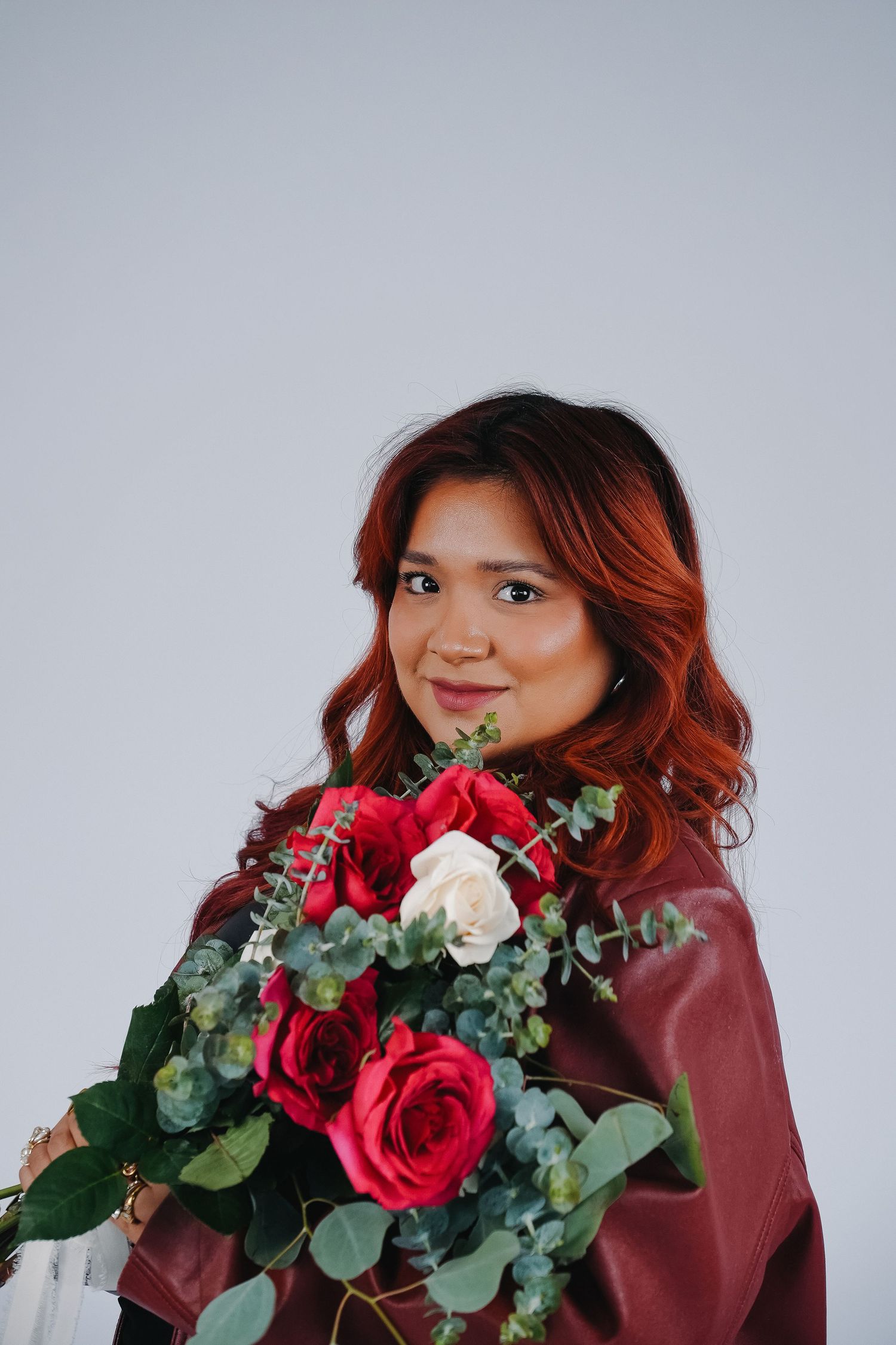 Woman with red hair holding a bouquet of red and white roses with eucalyptus, wearing a burgundy jacket.