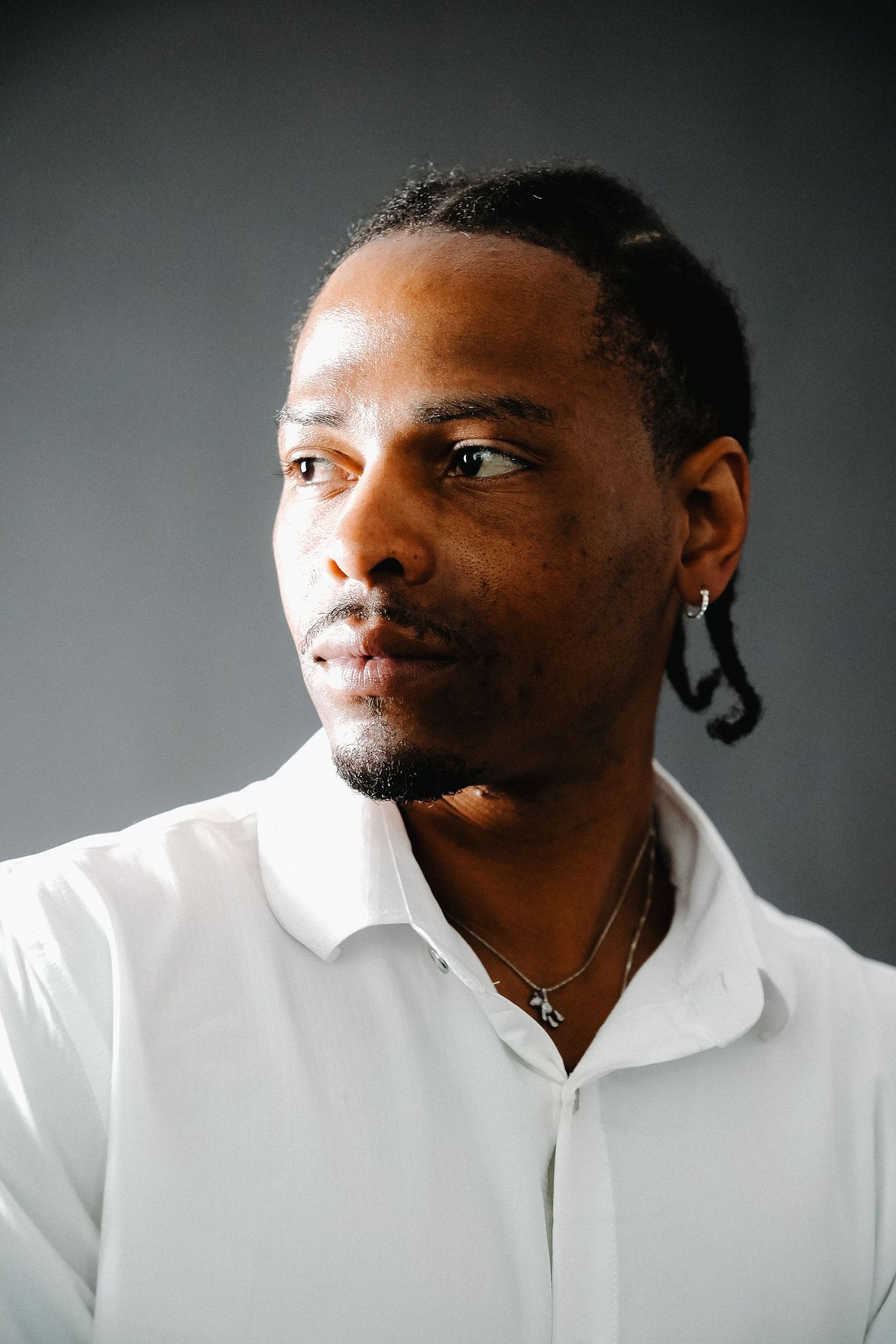 Young Black man in a white shirt with braided hair and earring looking away against a gray background.