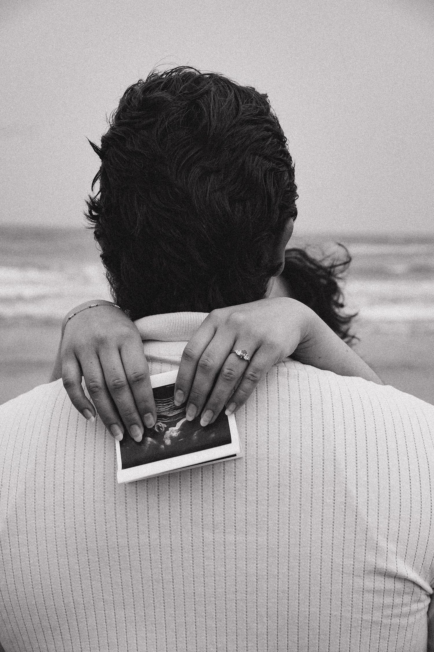 Black and white photo of woman's hands holding ultrasound on man's back during beach maternity session.