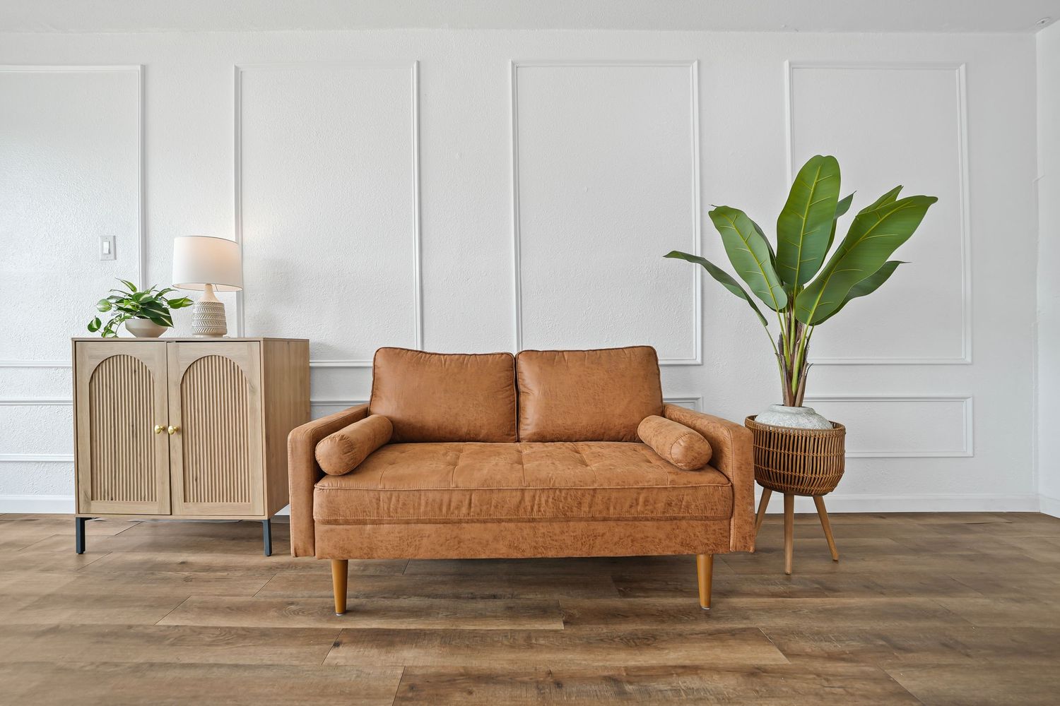 Clean modern living room with tan leather sofa, wooden cabinet, large tropical plant, and white panel walls.