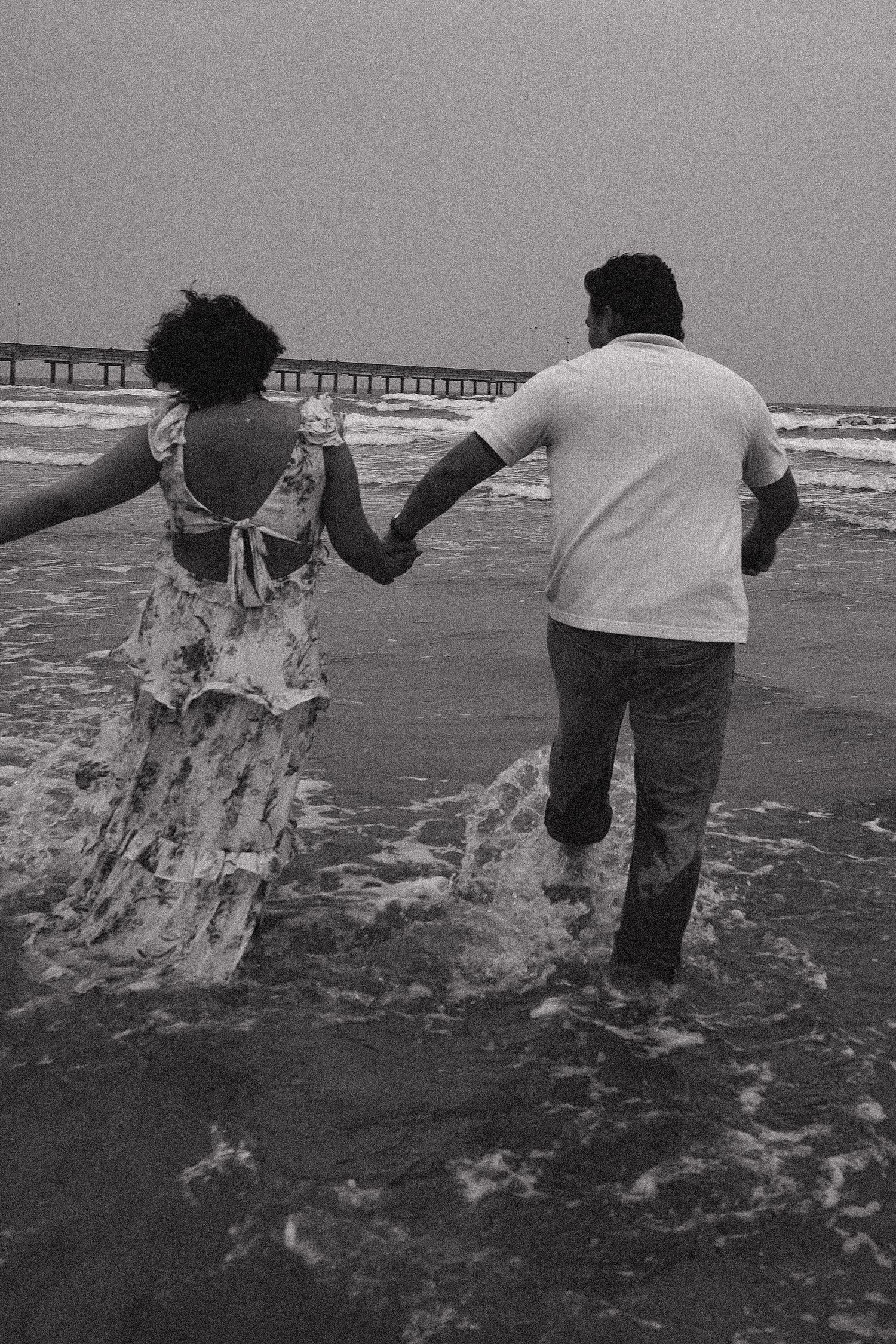 Couple holding hands running through ocean waves on beach in black and white photo.