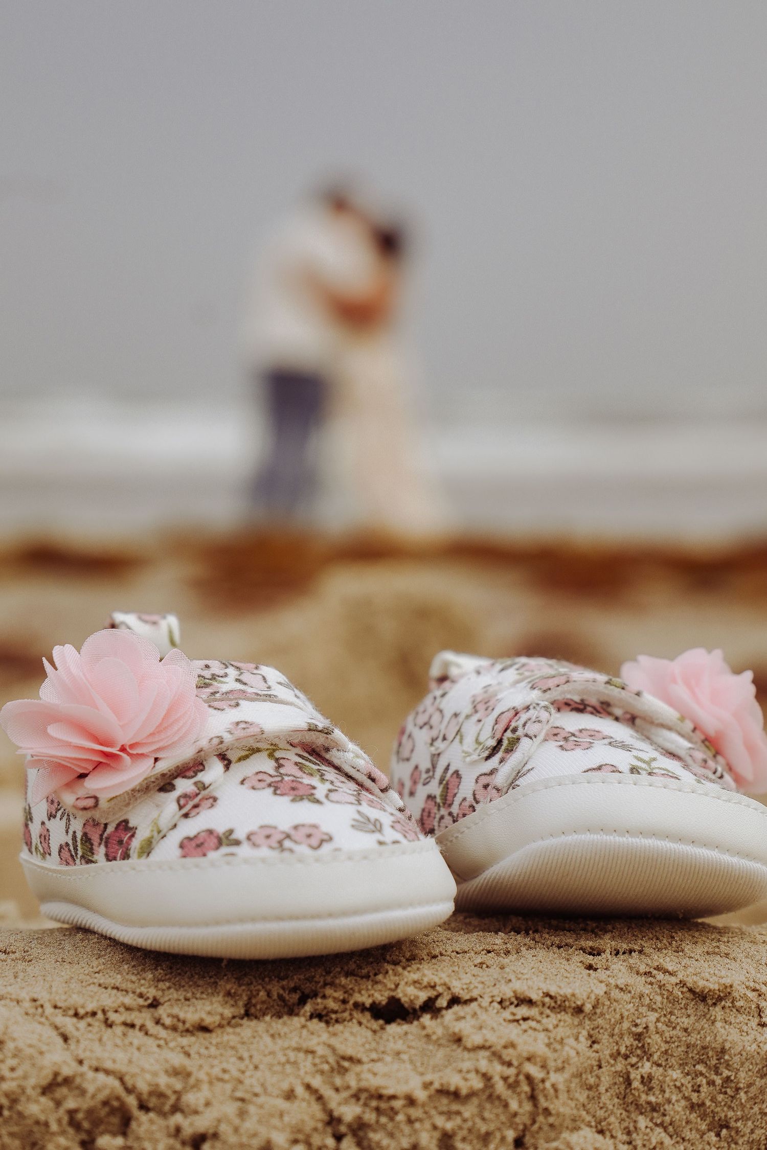 Floral baby shoes with pink flowers in sand with couple embracing at ocean beach in soft focus background.