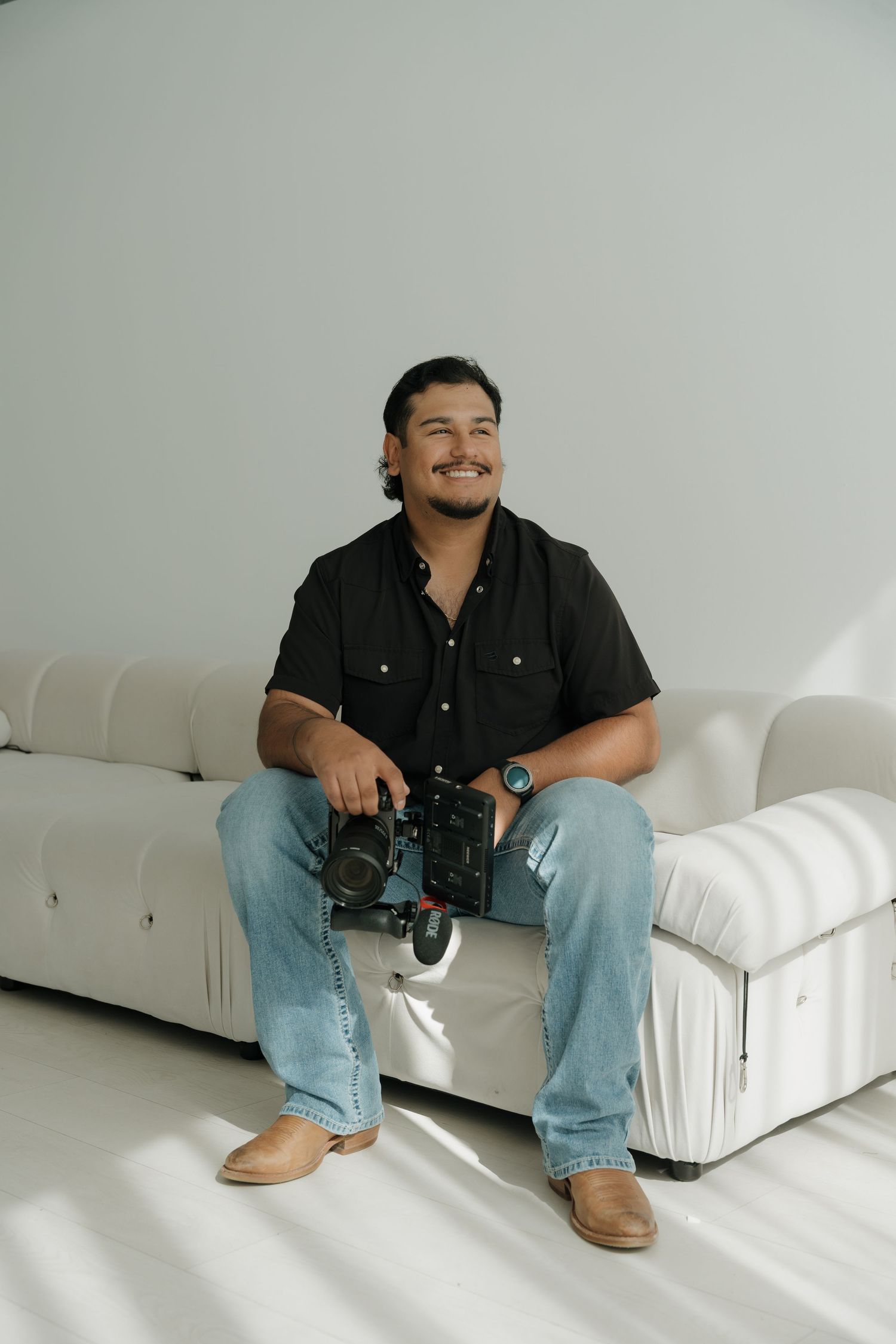 Smiling man in black shirt holding camera equipment while sitting on white couch in bright studio.