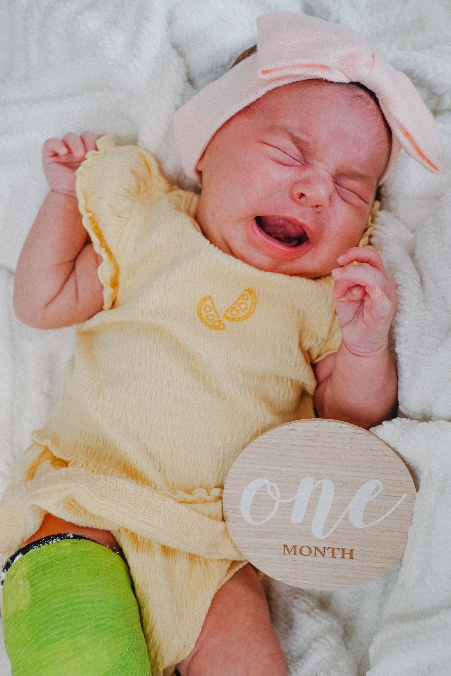 Crying one month old baby girl in yellow onesie and pink bow headband lying on white blanket with milestone disc.