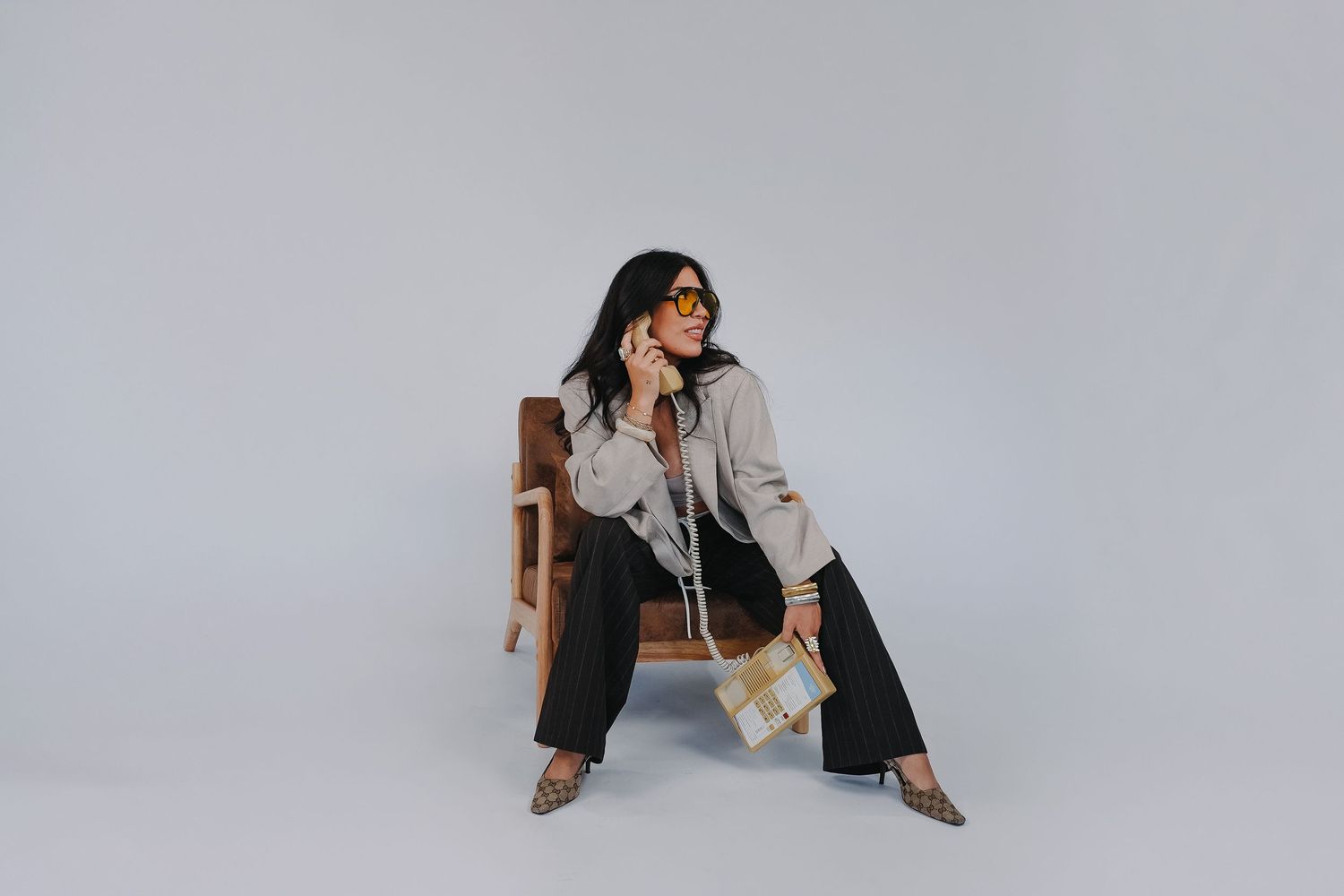 Woman in beige jacket, black wide-leg pants, sunglasses, and woven bag seated on wooden chair against gray background.