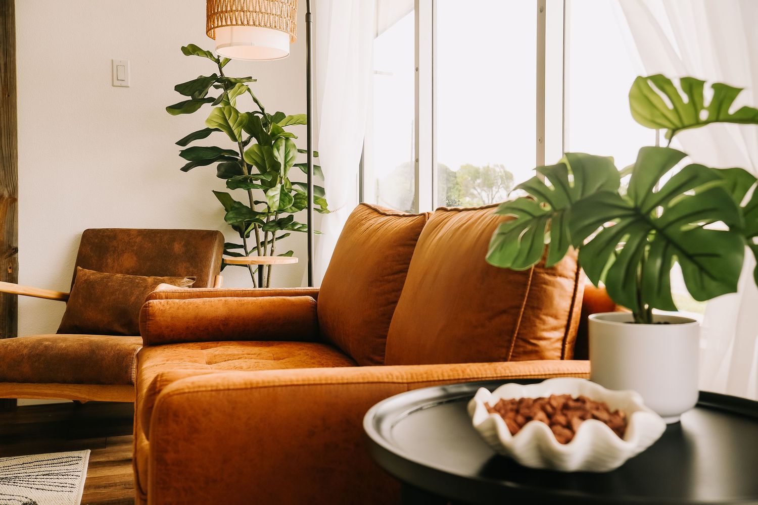 Warm brown leather sofa with monstera plant and decorative tray on dark coffee table in bright room.