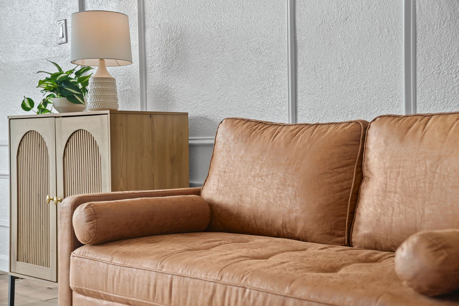 Close-up of tan leather loveseat beside a wooden arched cabinet with lamp and small green plant.