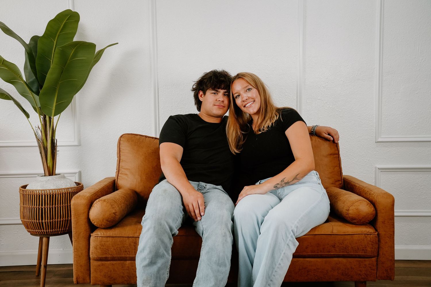 Couple sitting together on a brown leather sofa, smiling, wearing black shirts and light jeans, with a plant nearby.