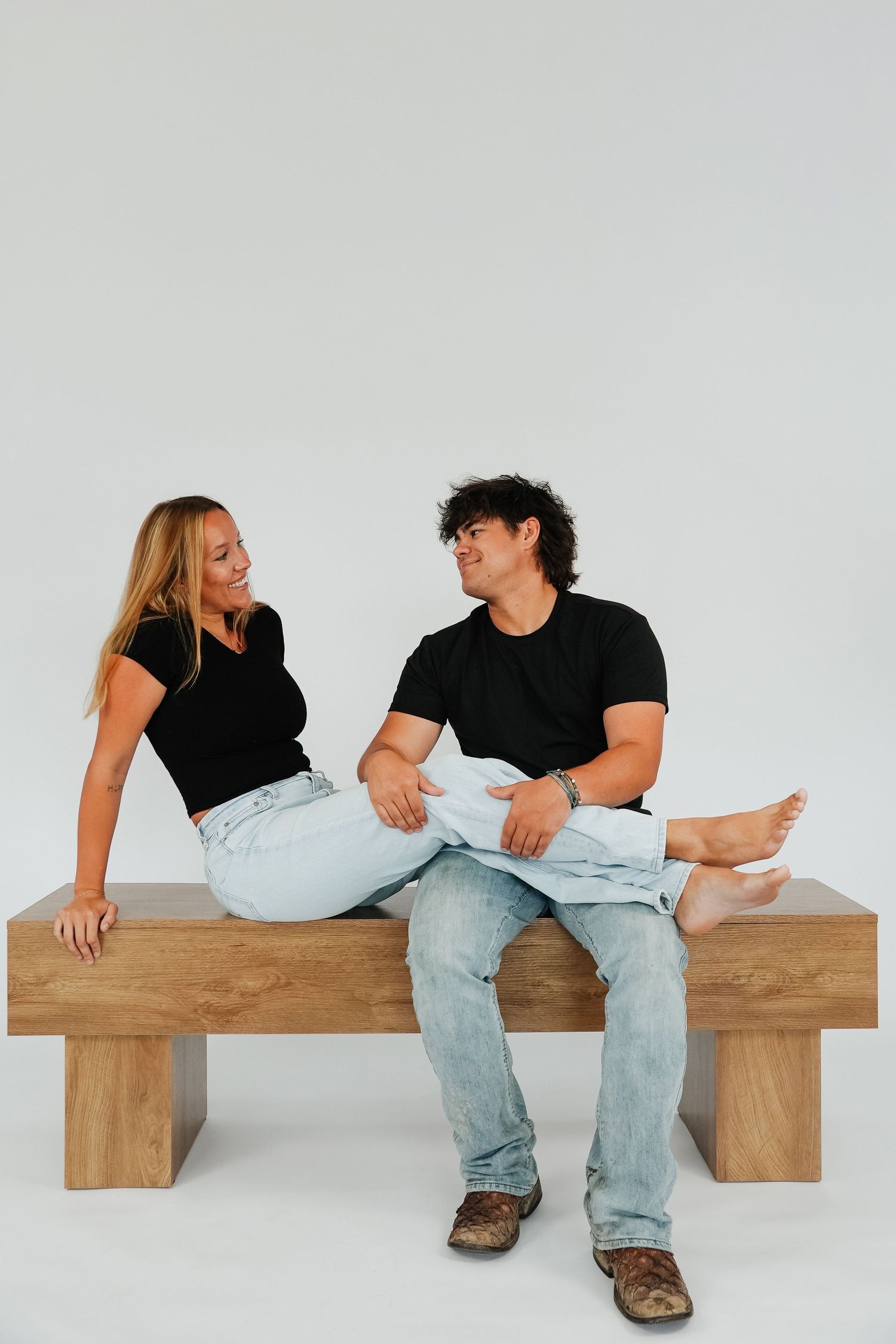 A couple in black shirts and jeans sit on a wooden bench, smiling at each other against a white background.