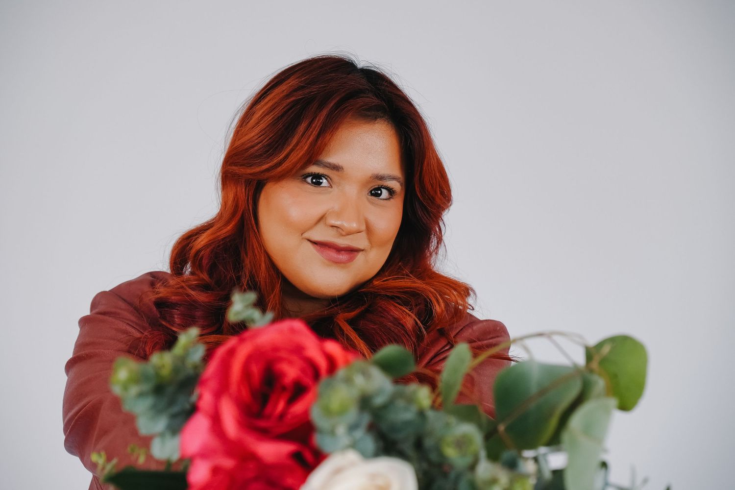 Smiling woman with red hair in a brown leather jacket posing with a bouquet of red and white roses.