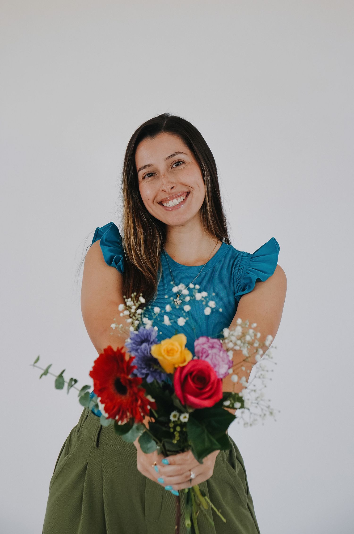 Smiling woman in teal top holding colorful bouquet of roses and wildflowers against white background.