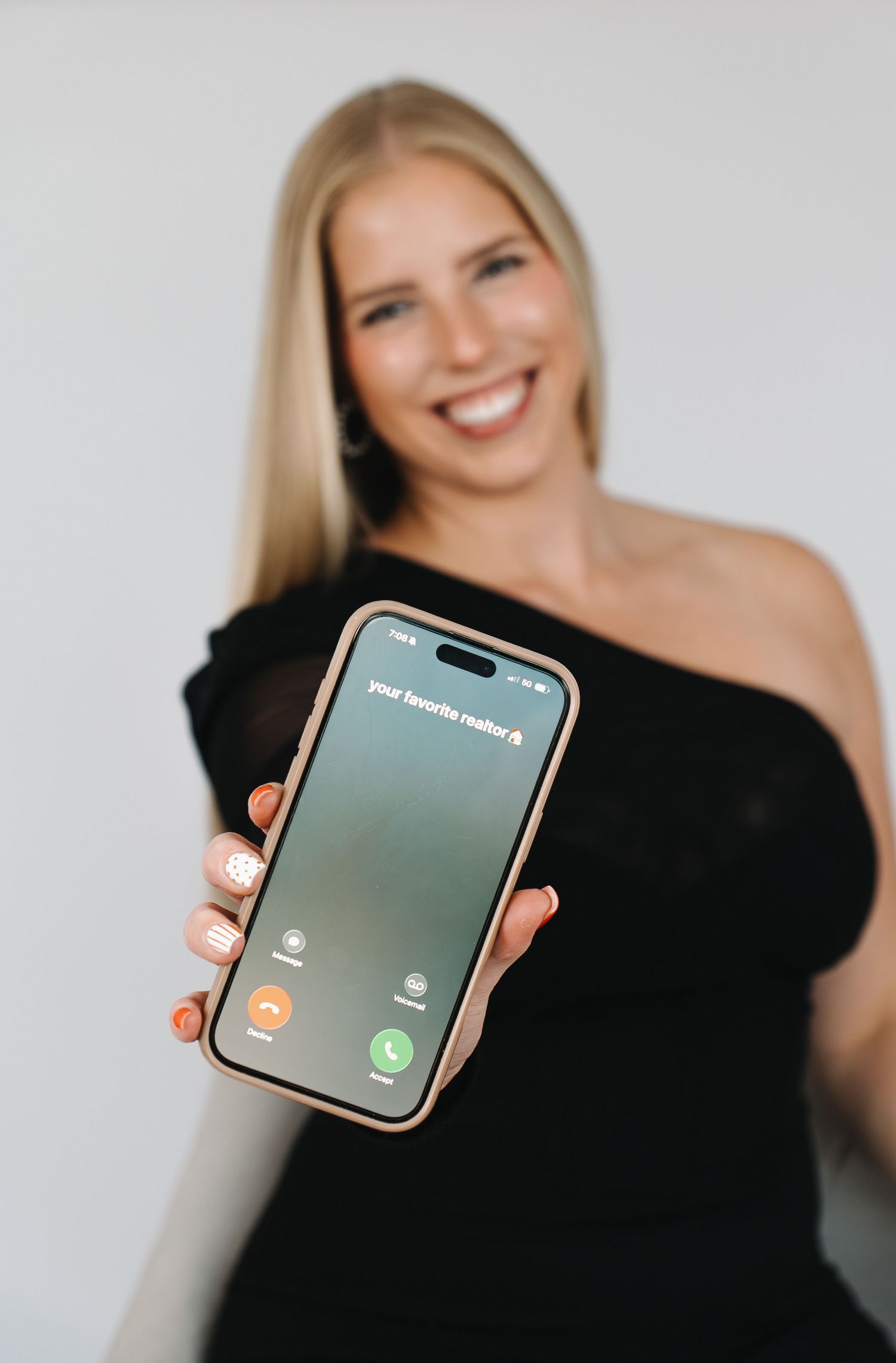 Smiling blonde woman in black off-shoulder top holding iPhone with incoming call screen toward camera.