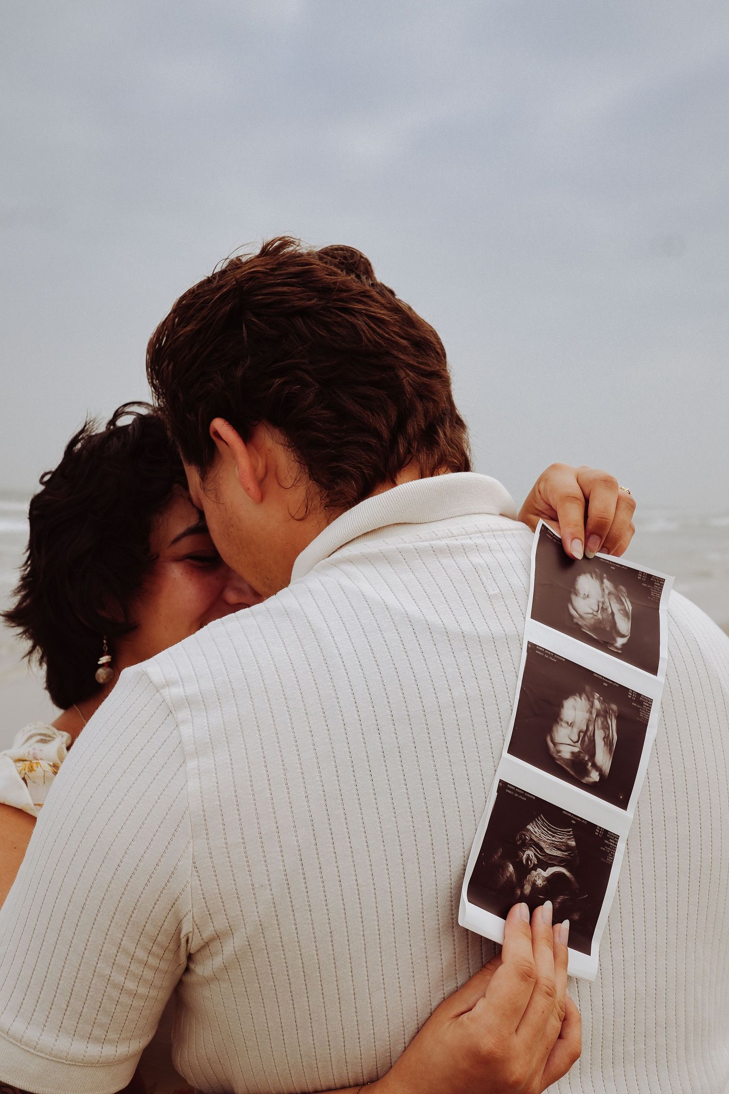 Couple embracing and kissing at beach while man holds ultrasound photos, viewed from behind near ocean waves.