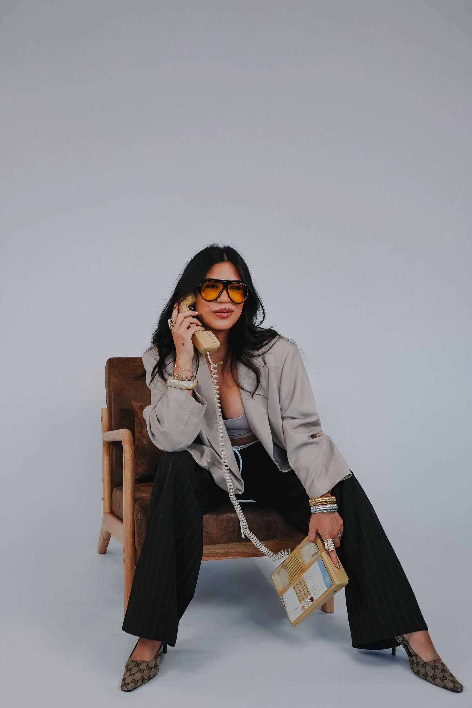 Woman in sunglasses sitting in chair holding vintage phone and designer bag, wearing jacket and wide-leg pants.