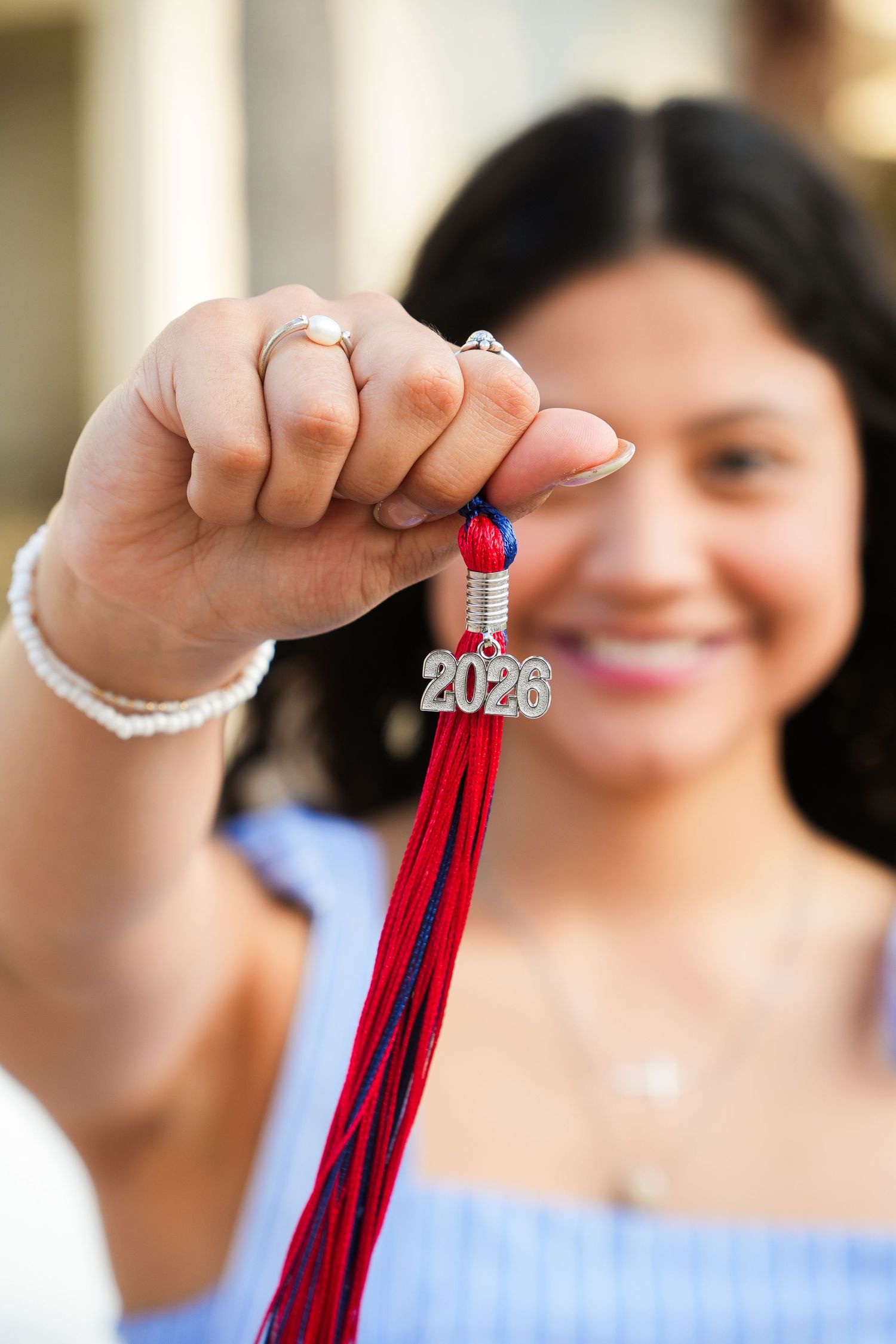 Smiling girl holds red and blue 2026 graduation tassel charm toward camera.