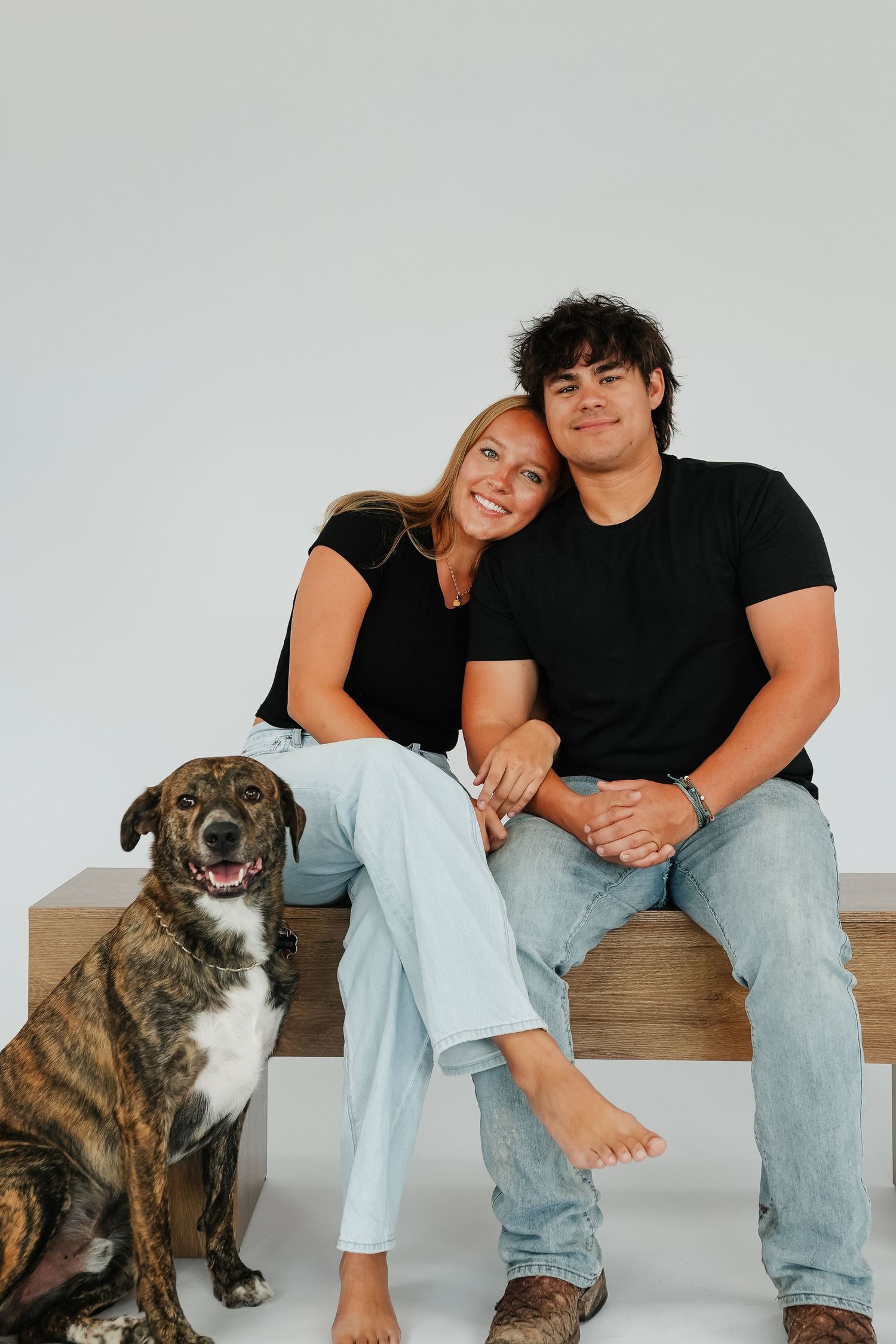 Smiling couple in black tees and jeans sitting on wooden bench with brindle dog against white background.