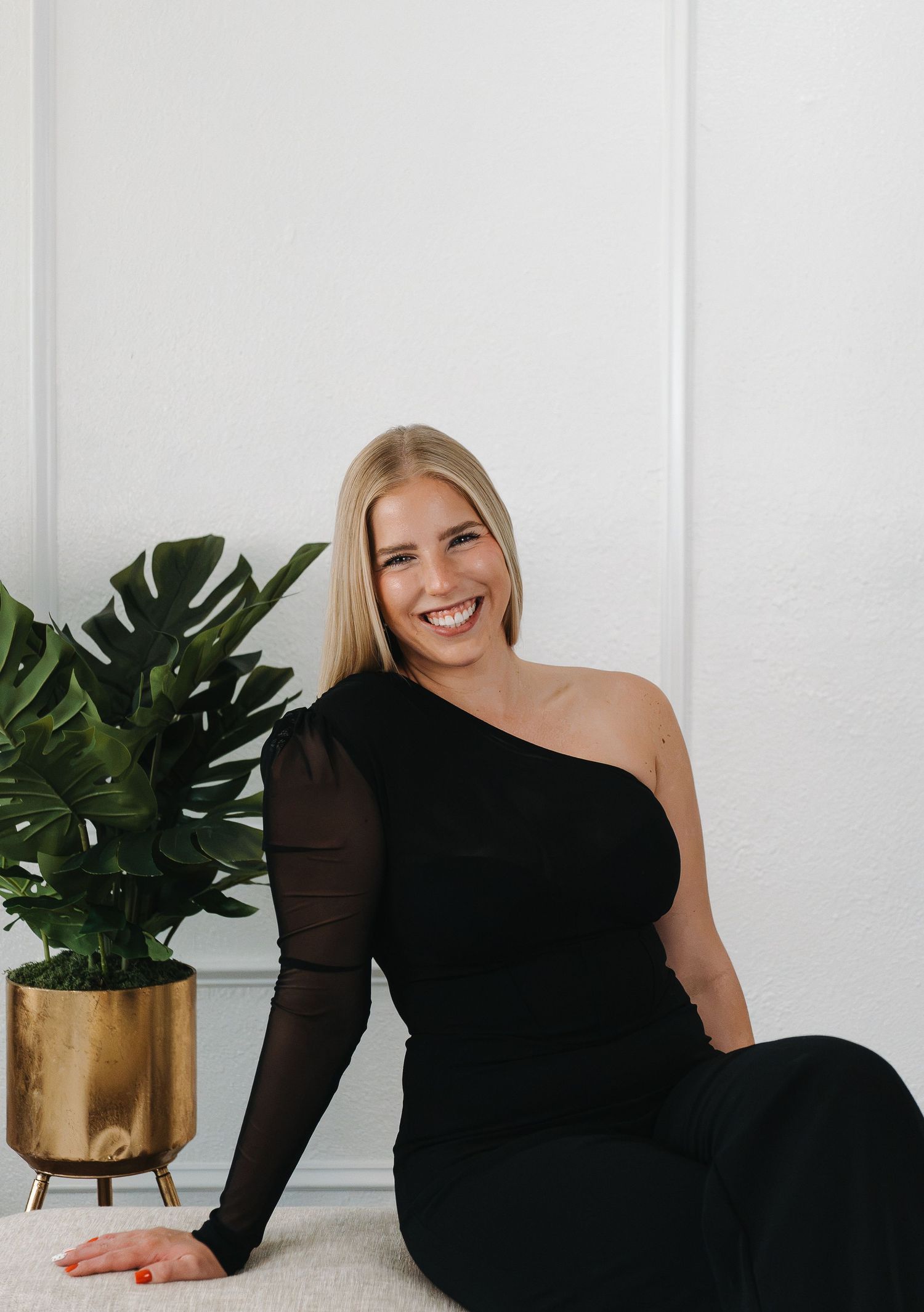 Smiling blonde woman in black one-shoulder outfit sitting on floor beside gold planter with monstera plant.