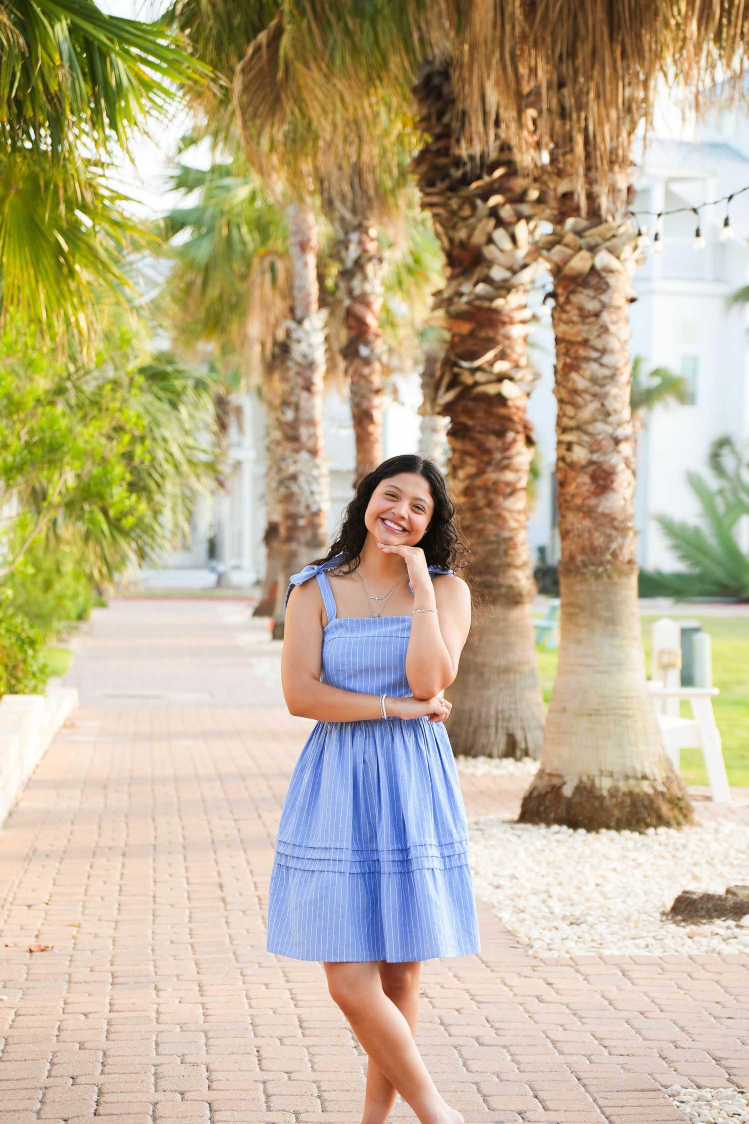 Young woman in blue sundress smiling among palm trees on brick walkway.
