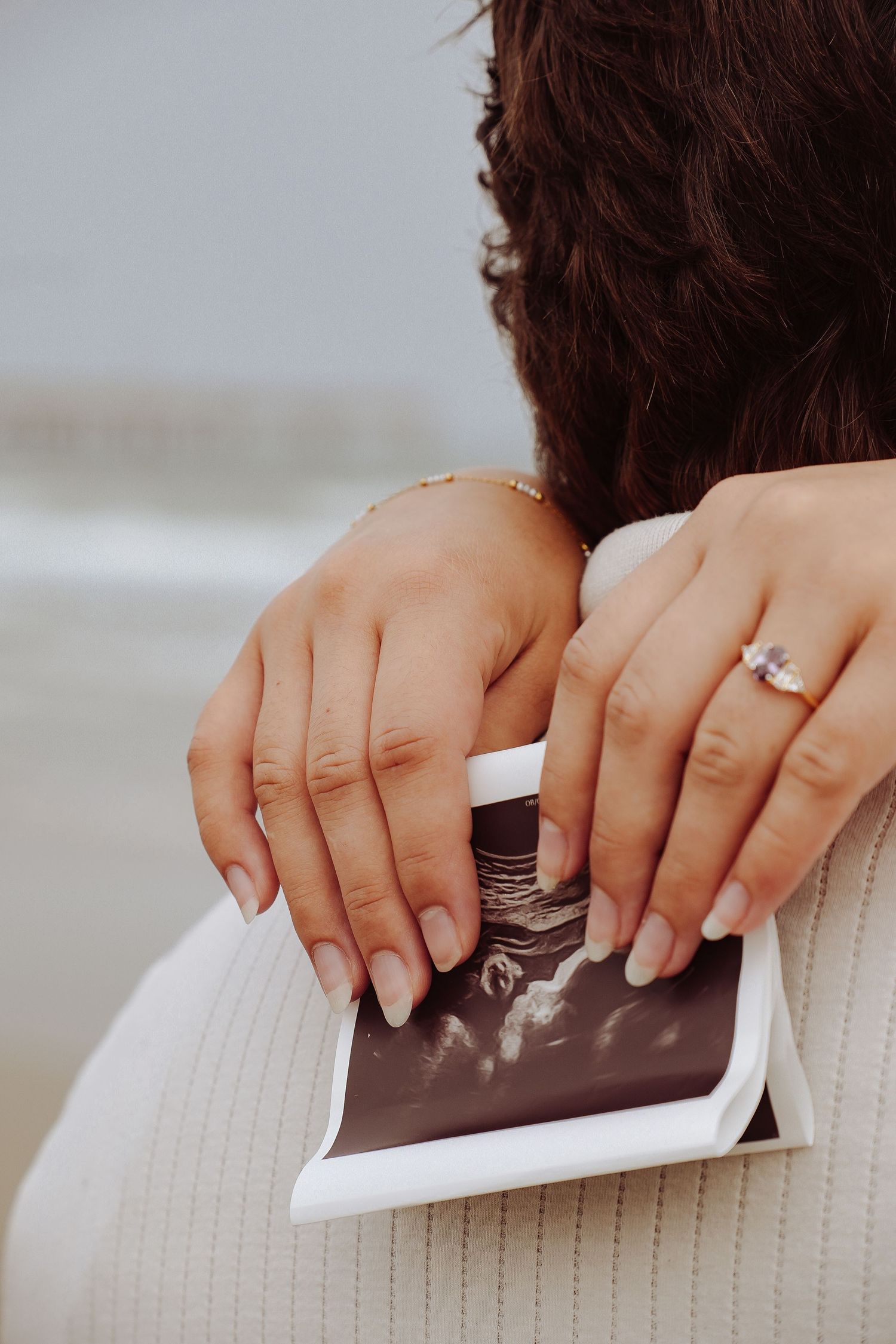Two hands holding ultrasound photos on shoulder, woman wearing diamond engagement ring at beach maternity shoot.