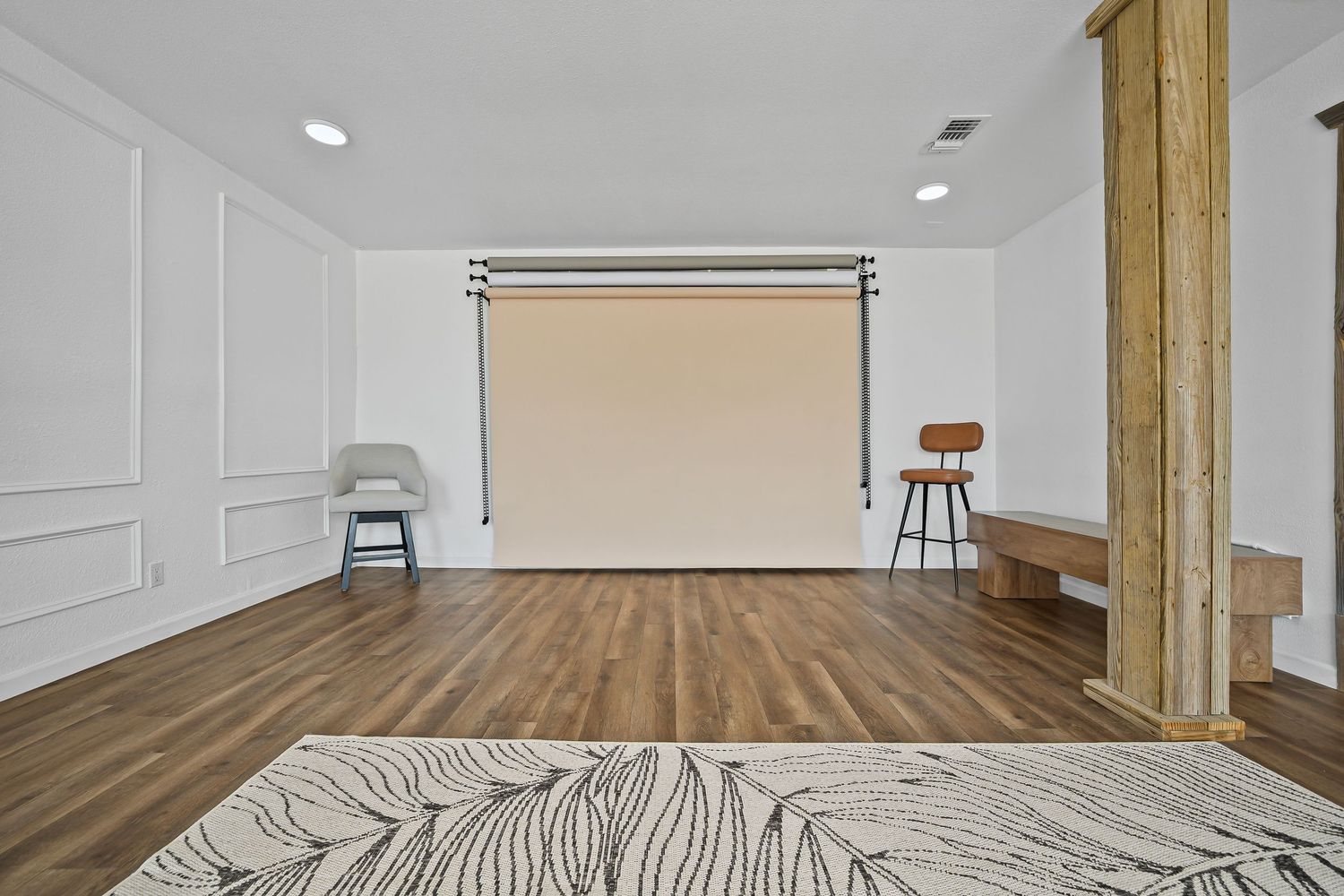 Open room with white wainscoting walls, wooden column, roller shade, hardwood floors, and minimalist decor.