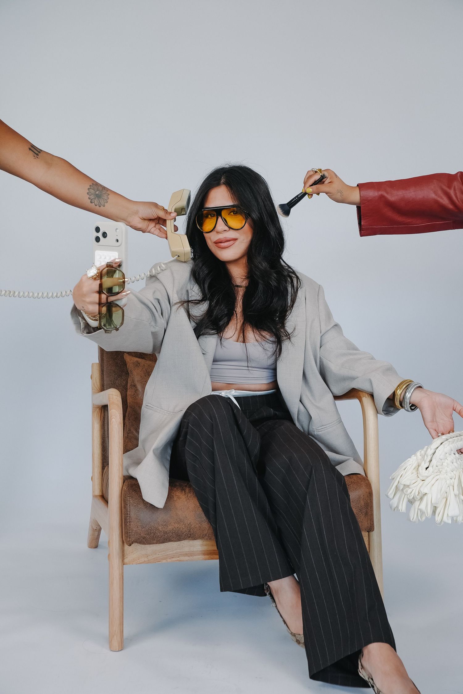 Woman in gray blazer and sunglasses sits in chair surrounded by hands holding phone, brush, and bag.