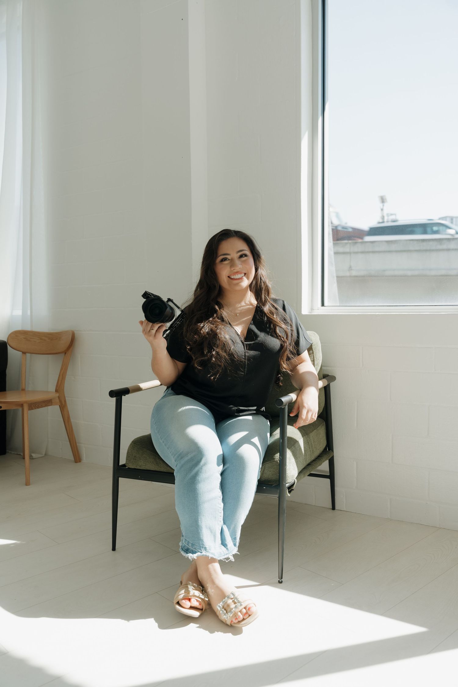 Smiling woman in black top and jeans sitting in chair holding camera near bright studio window.