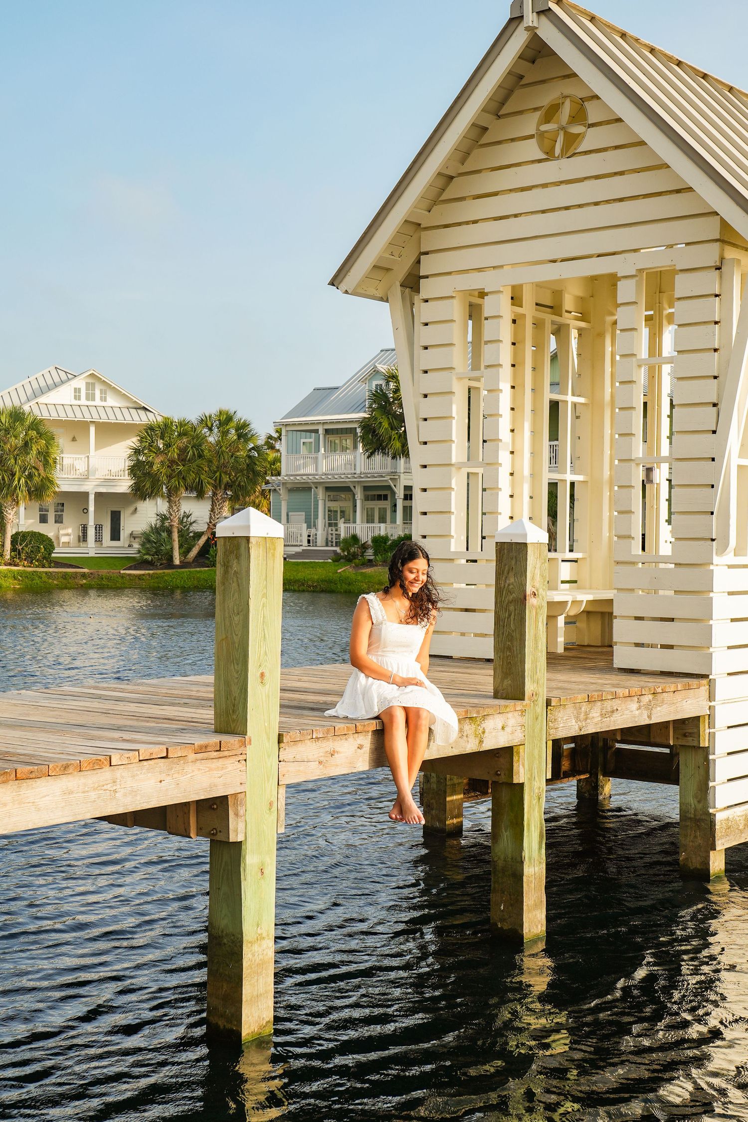 Young woman in white dress sits on wooden dock edge beside a charming white boathouse over calm water.