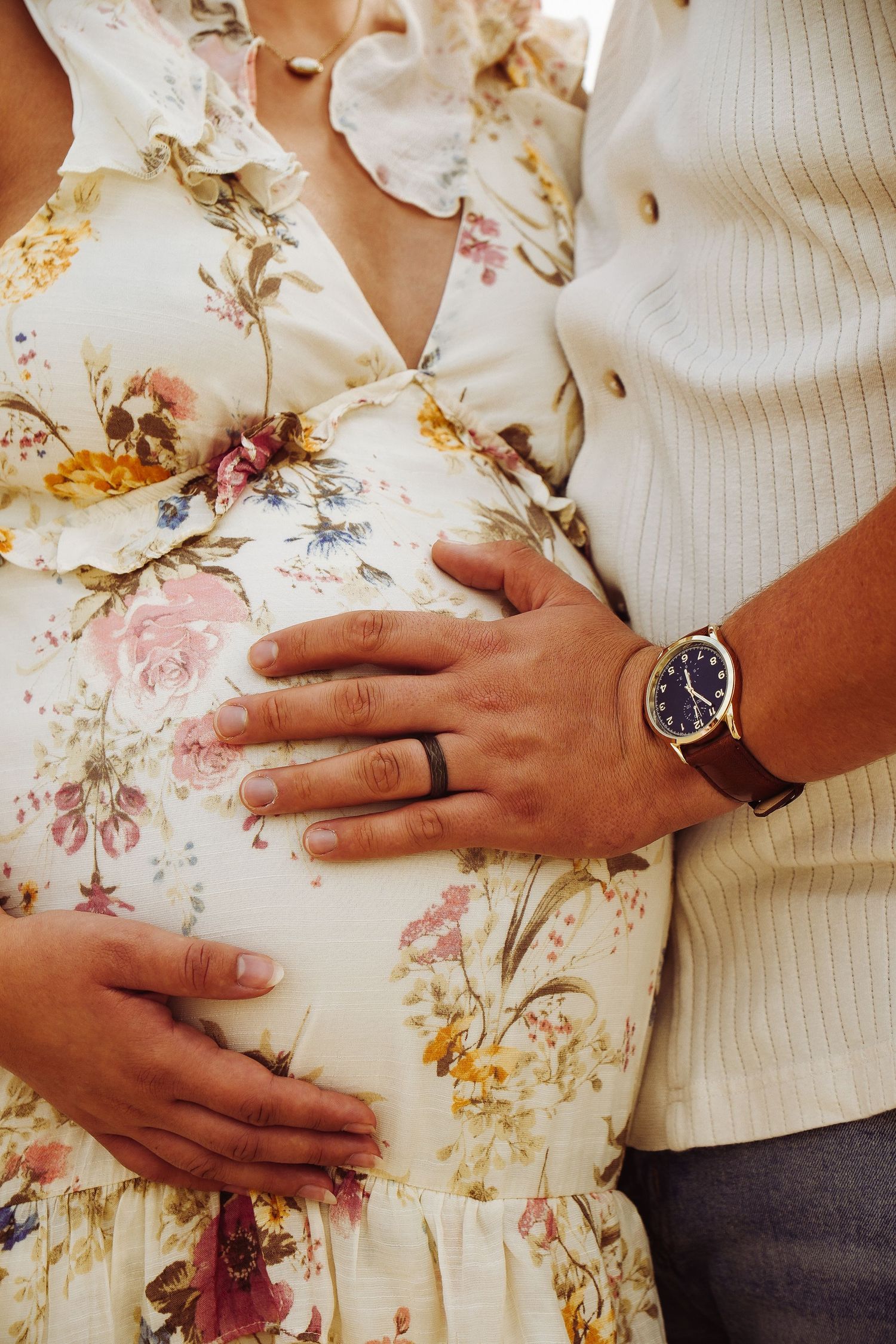 Couple cradling a pregnant belly, wearing a floral maternity dress and a navy watch, in a tender pose.