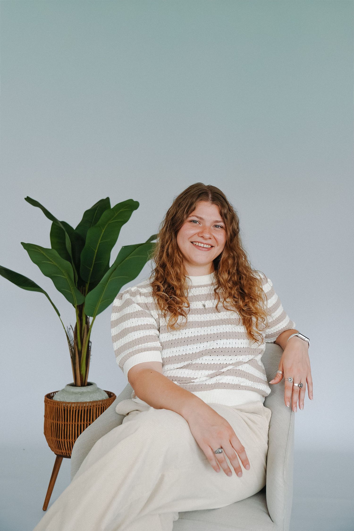 Smiling woman with curly hair sitting in a gray chair wearing a striped sweater beside a tropical plant.