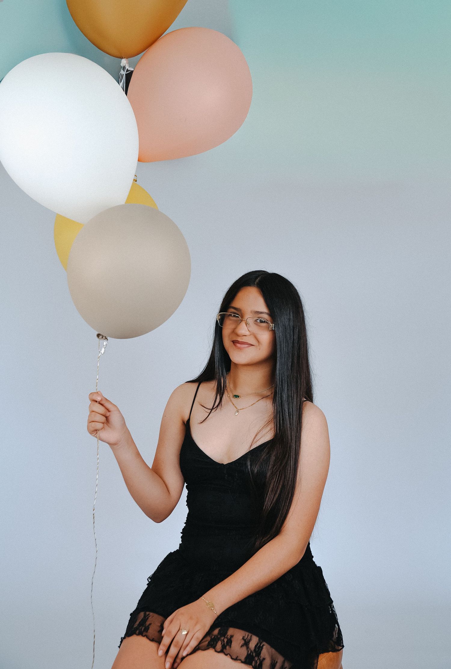 Young woman in black lace dress holding colorful balloons, smiling against a light blue background.