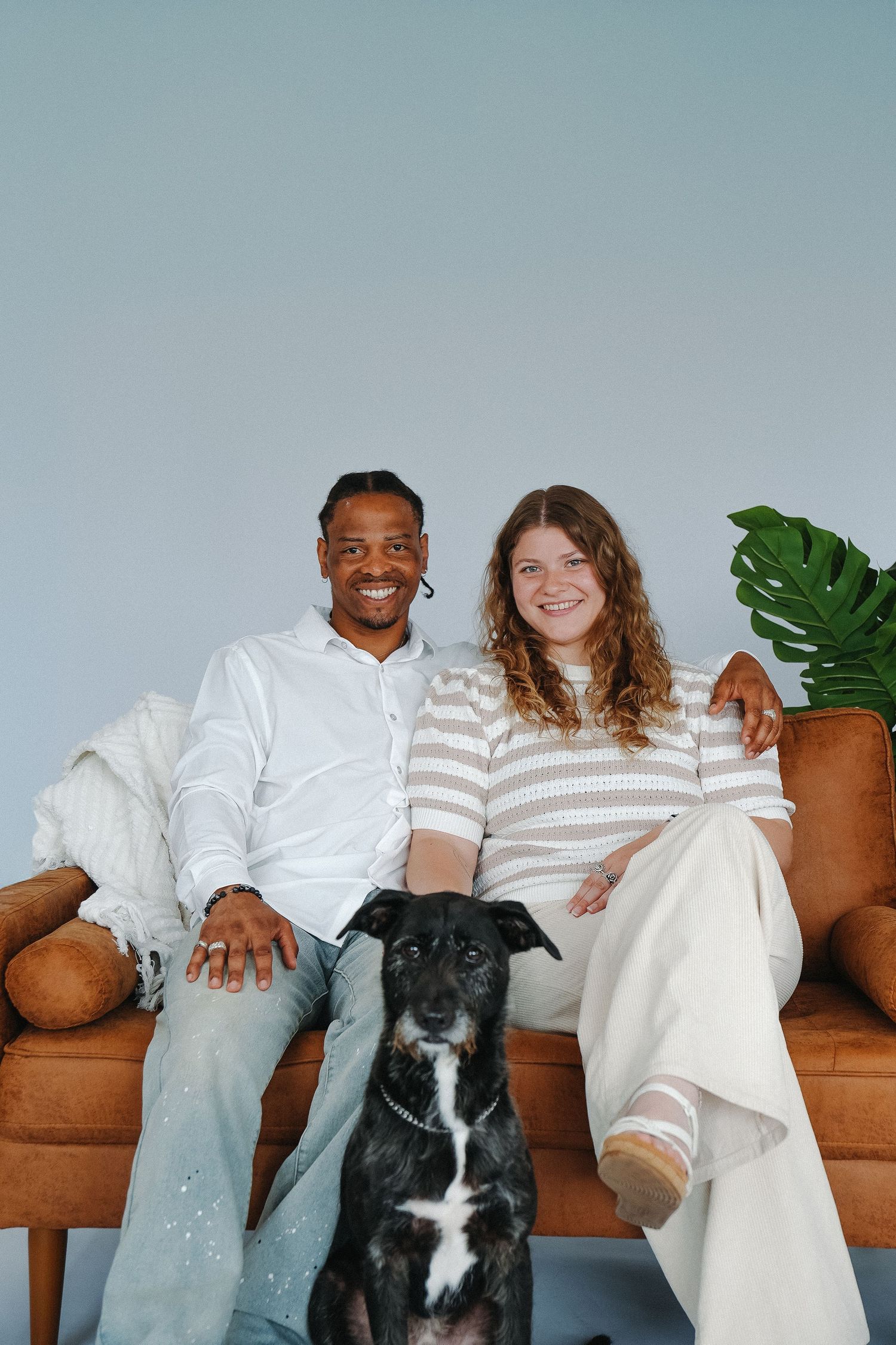 A smiling interracial couple sits on a brown leather sofa with their black and white dog in a bright modern living room.