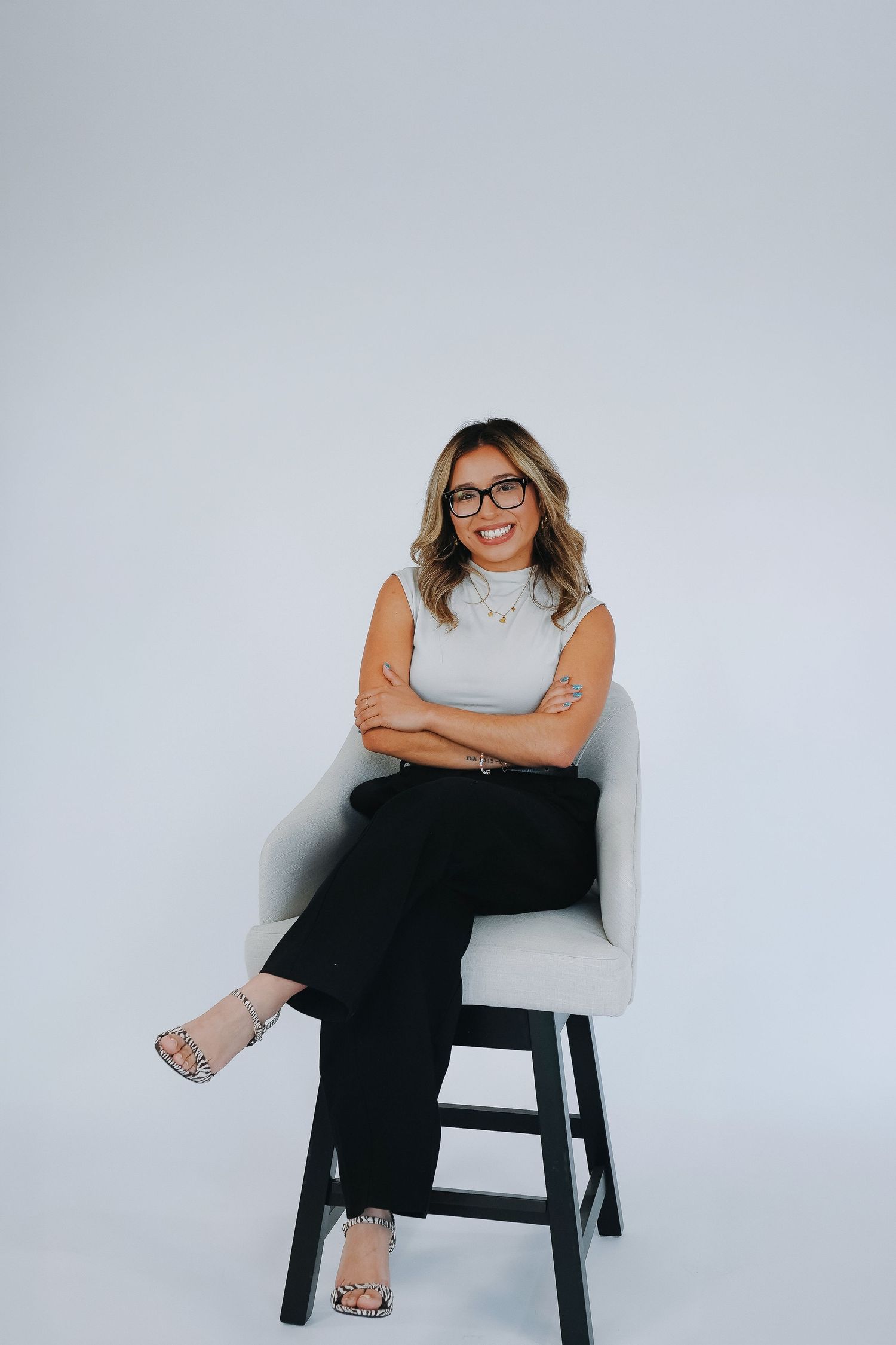 Professional woman in glasses sits cross-legged on chair, wearing white top and black pants, smiling confidently.