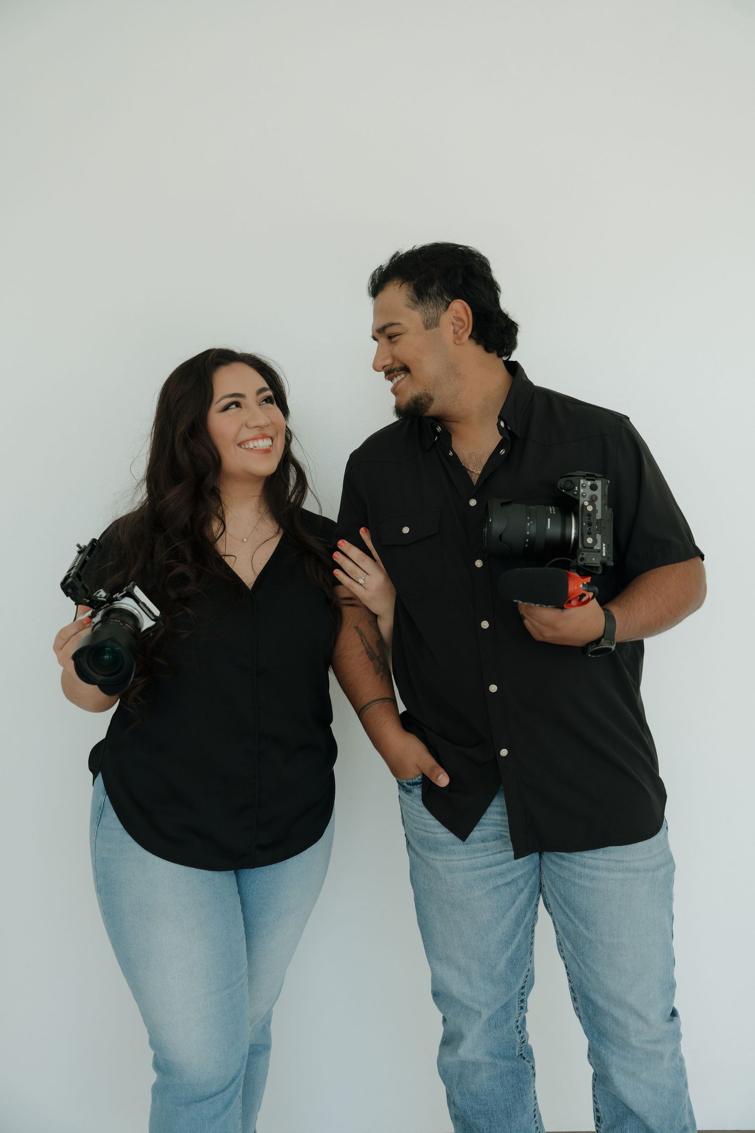 Two photographers in black shirts and jeans smiling and holding cameras against white background.