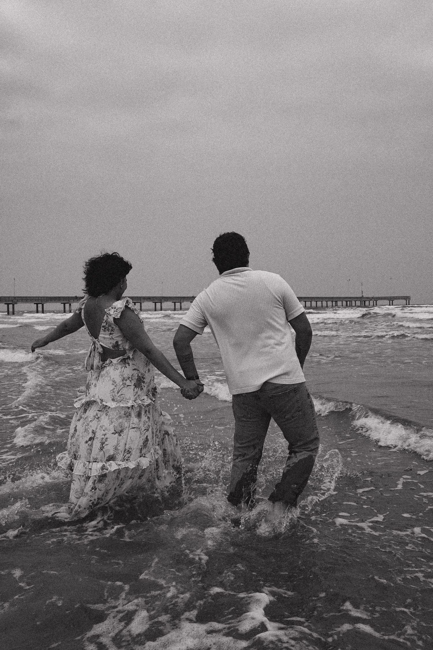 Black and white photo of a couple holding hands while wading through ocean waves with a pier in the distance.