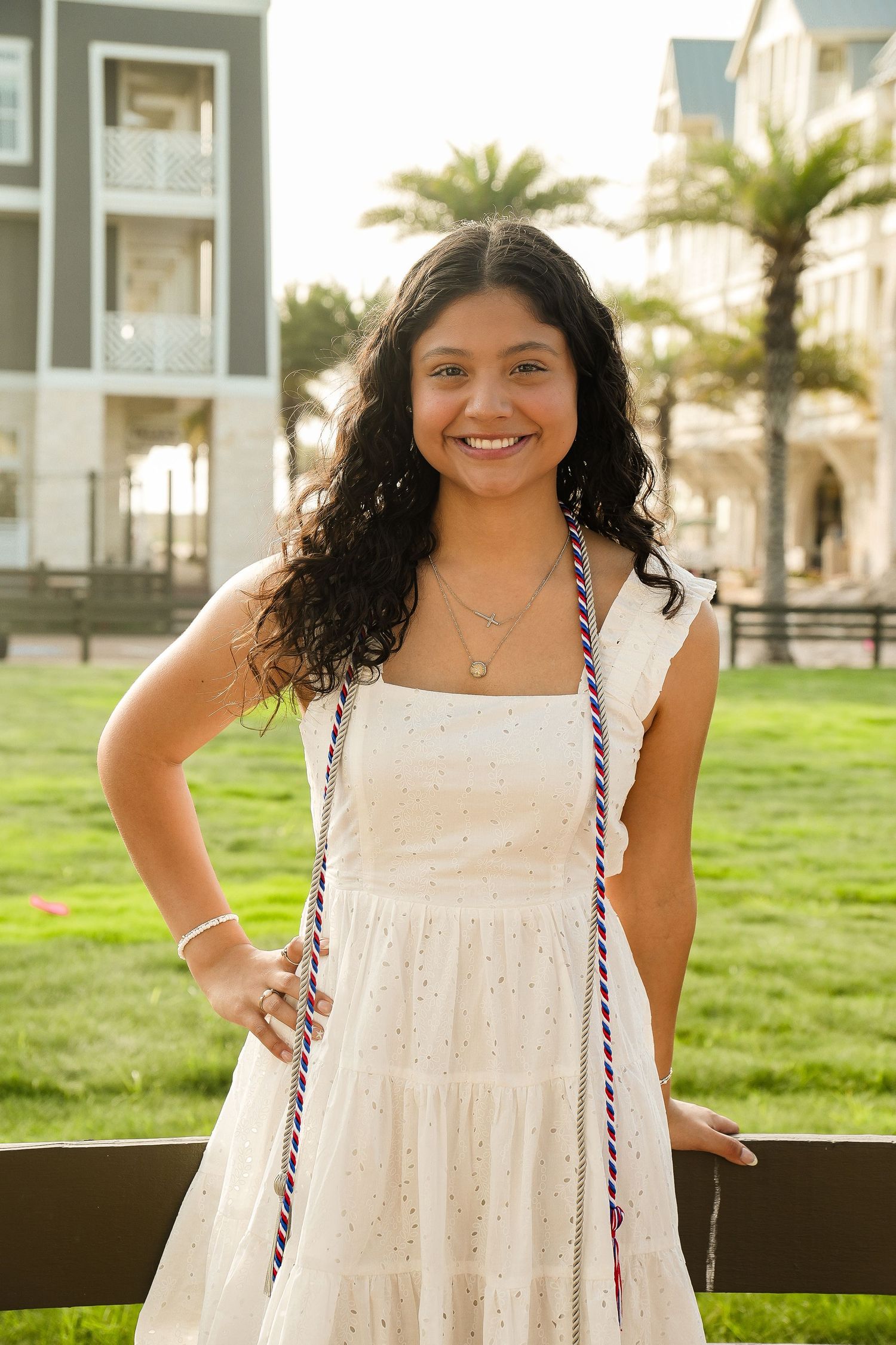 Smiling young woman in white dress with graduation cords poses outdoors near coastal buildings and palm trees.