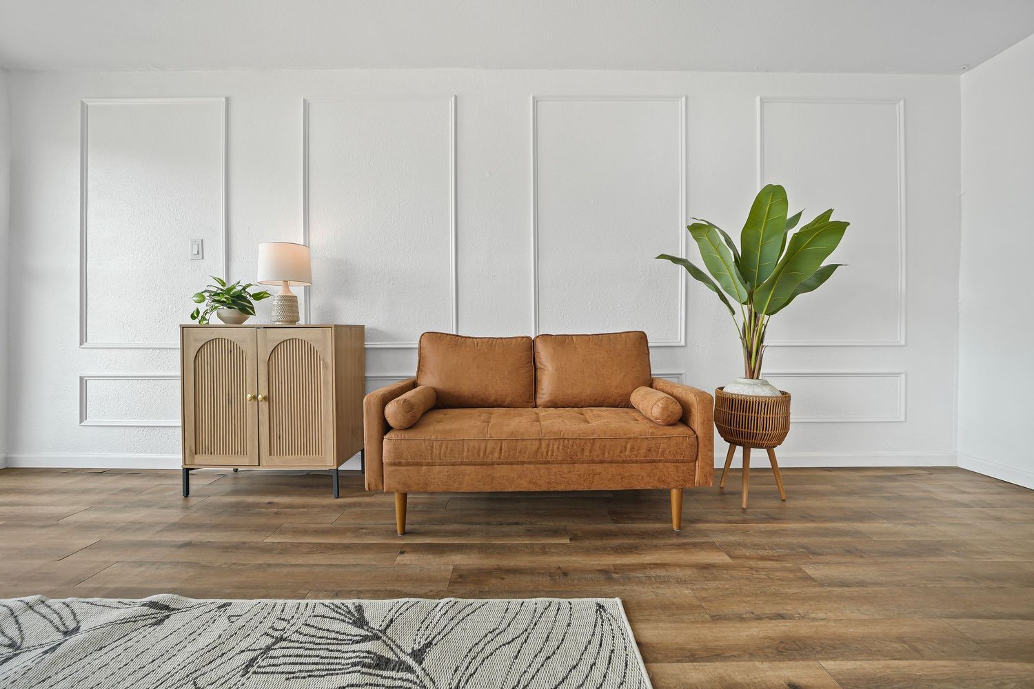 Minimalist living room with tan leather loveseat, arched wood cabinet, tall tropical plant, and white wainscoting.