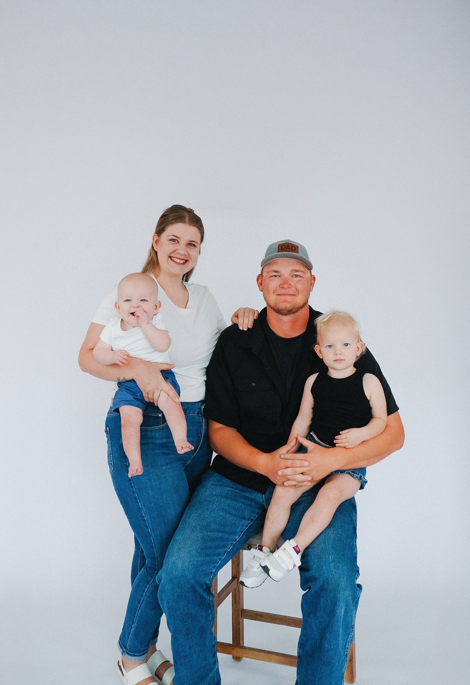 Young family portrait with mother, father, and two toddlers posing together against a white background.