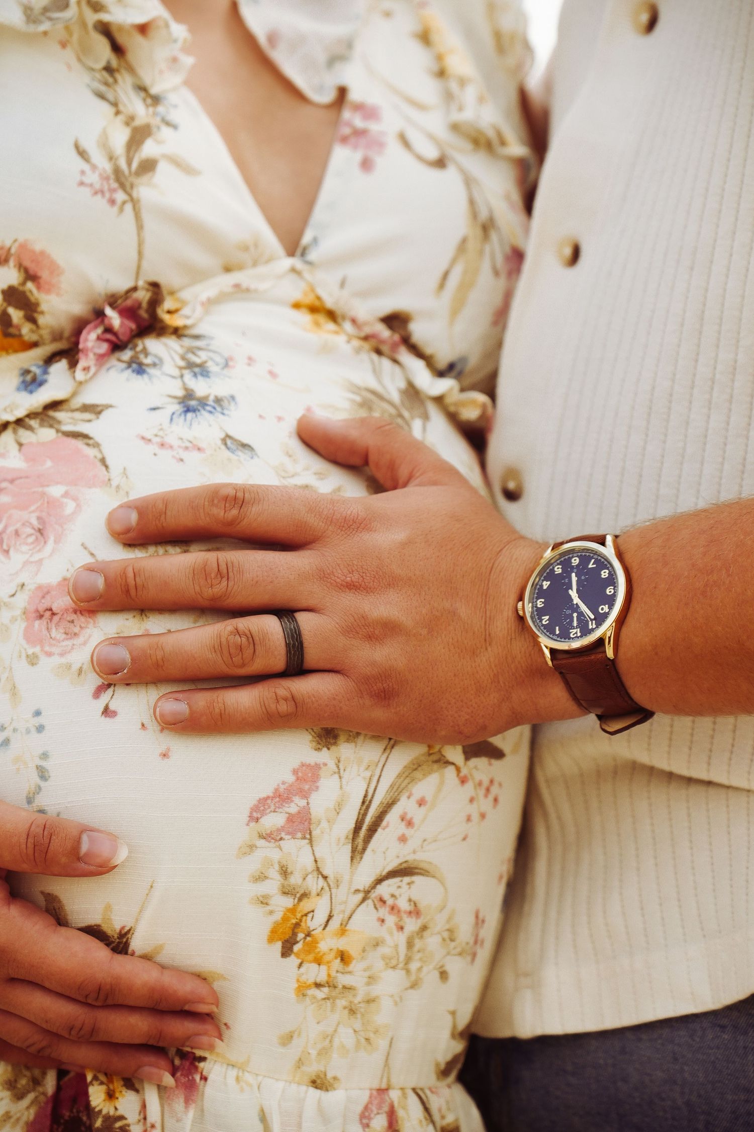 Close-up of pregnant belly with couple's hands, woman wearing black silicone ring and man wearing gold watch.