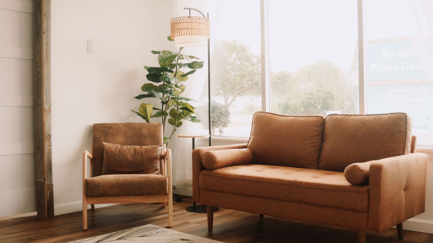 Modern living room with tan leather loveseat, matching armchair, floor lamp, and fiddle leaf fig plant near large windows.