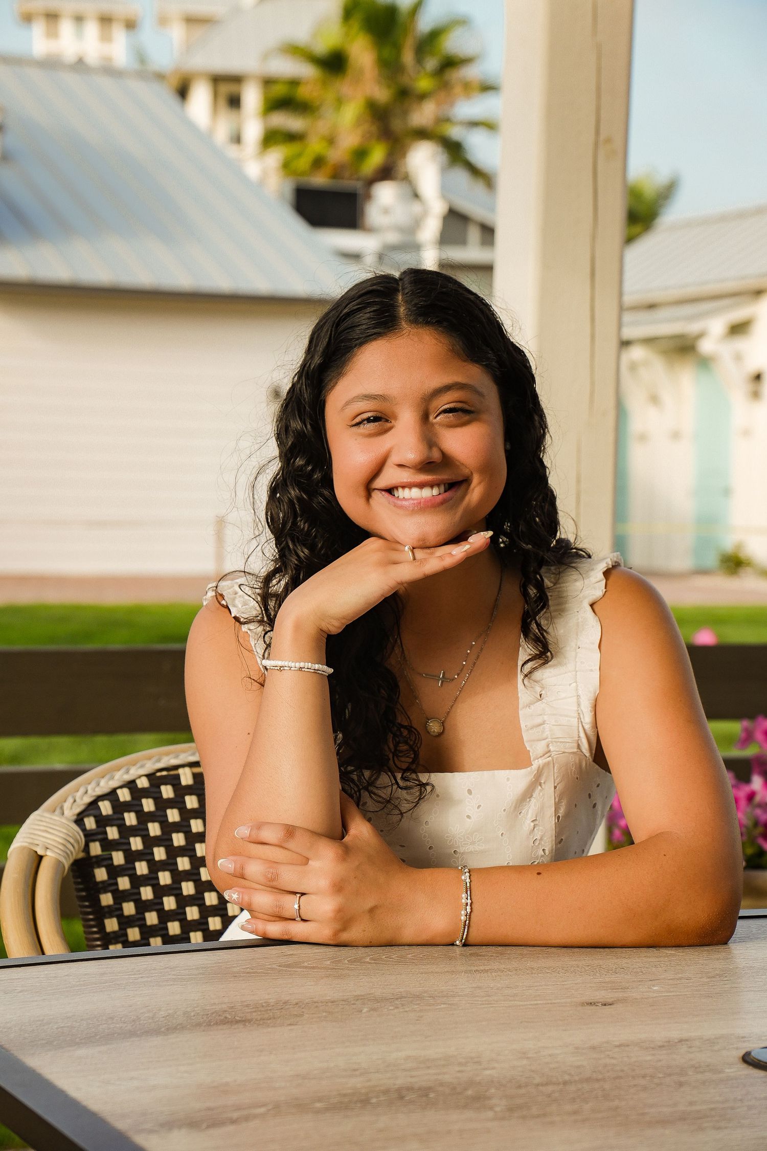 Smiling young woman in white dress sits at outdoor table with chin resting on hands near coastal buildings.