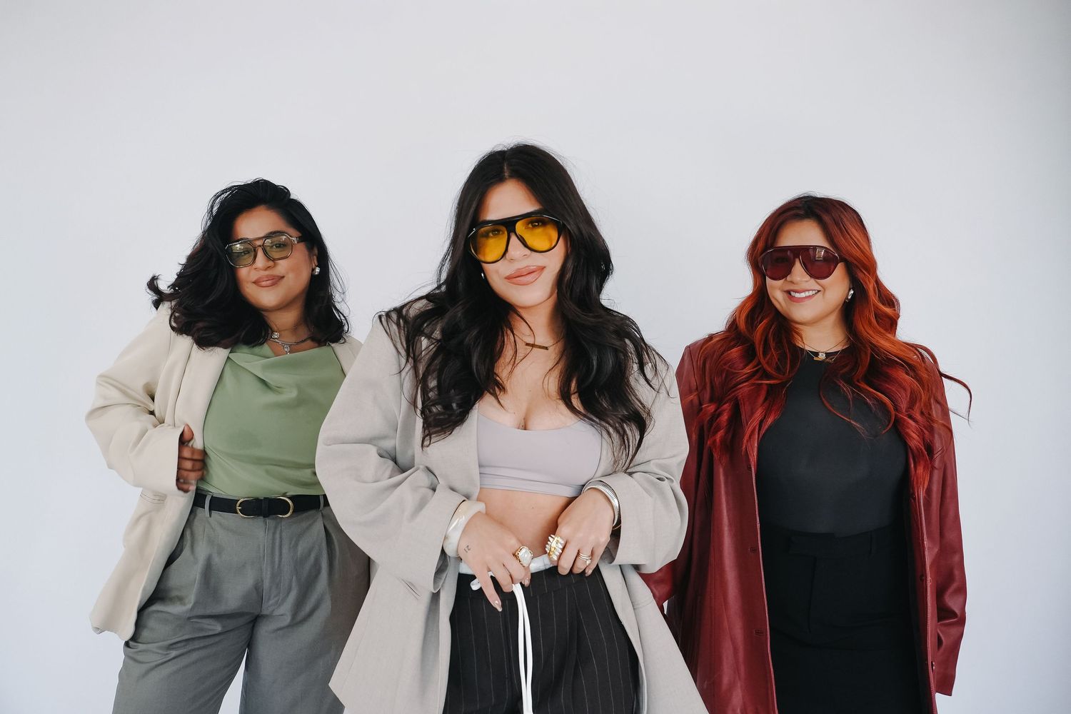 Three stylish women wearing sunglasses pose together against a white background in trendy outfits.
